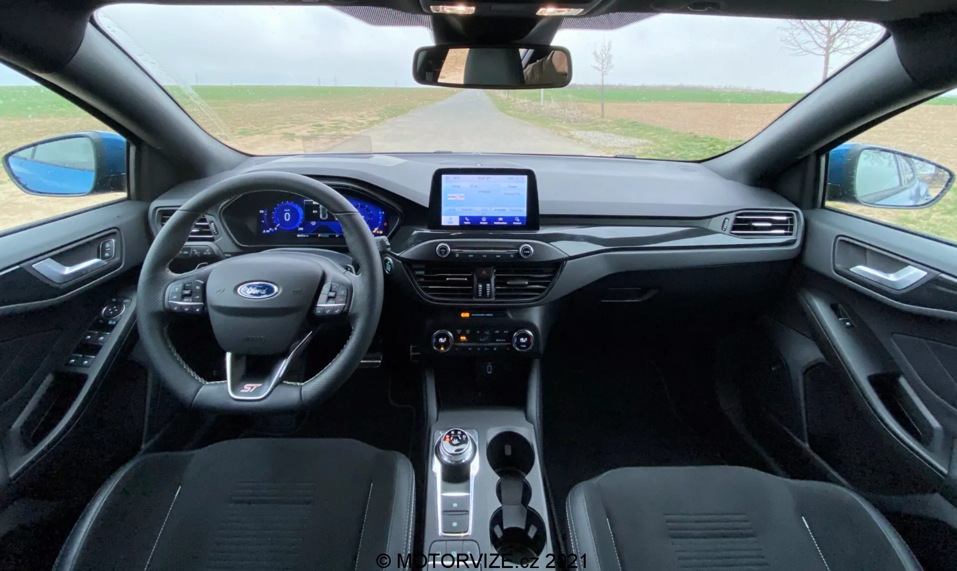 Vista interna della Ford Focus 2022 dall'area dei sedili posteriori, evidenziando il moderno volante multifunzione con comandi integrati, quadro strumenti completamente digitale, sistema di infotainment touchscreen, pulsanti di controllo del clima lucidi, leva del cambio rotante, sedili anteriori sagomati rivestiti in materiale scuro e pannelli porta abbinati con maniglie integrate e superfici di controllo per finestre e specchietti.