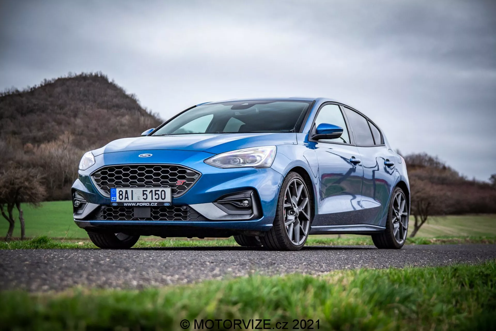 Una Ford Focus 2022 di colore blu chiaro posizionata su un terreno erboso con colline e un cielo tenue sullo sfondo. La foto cattura una vista anteriore a tre quarti, concentrandosi sul lato sinistro dell'auto. Le caratteristiche visibili includono una grande griglia anteriore a nido d'ape, un paraurti anteriore sportivo con fendinebbia, fari LED eleganti, cerchi in lega a più razze e specchietti laterali del colore della carrozzeria. L'auto ha una finitura con vernice metallizzata, aggiungendo al suo aspetto dinamico e sportivo. Lo sfondo è leggermente sfocato, sottolineando il design del veicolo e le caratteristiche orientate alle prestazioni.