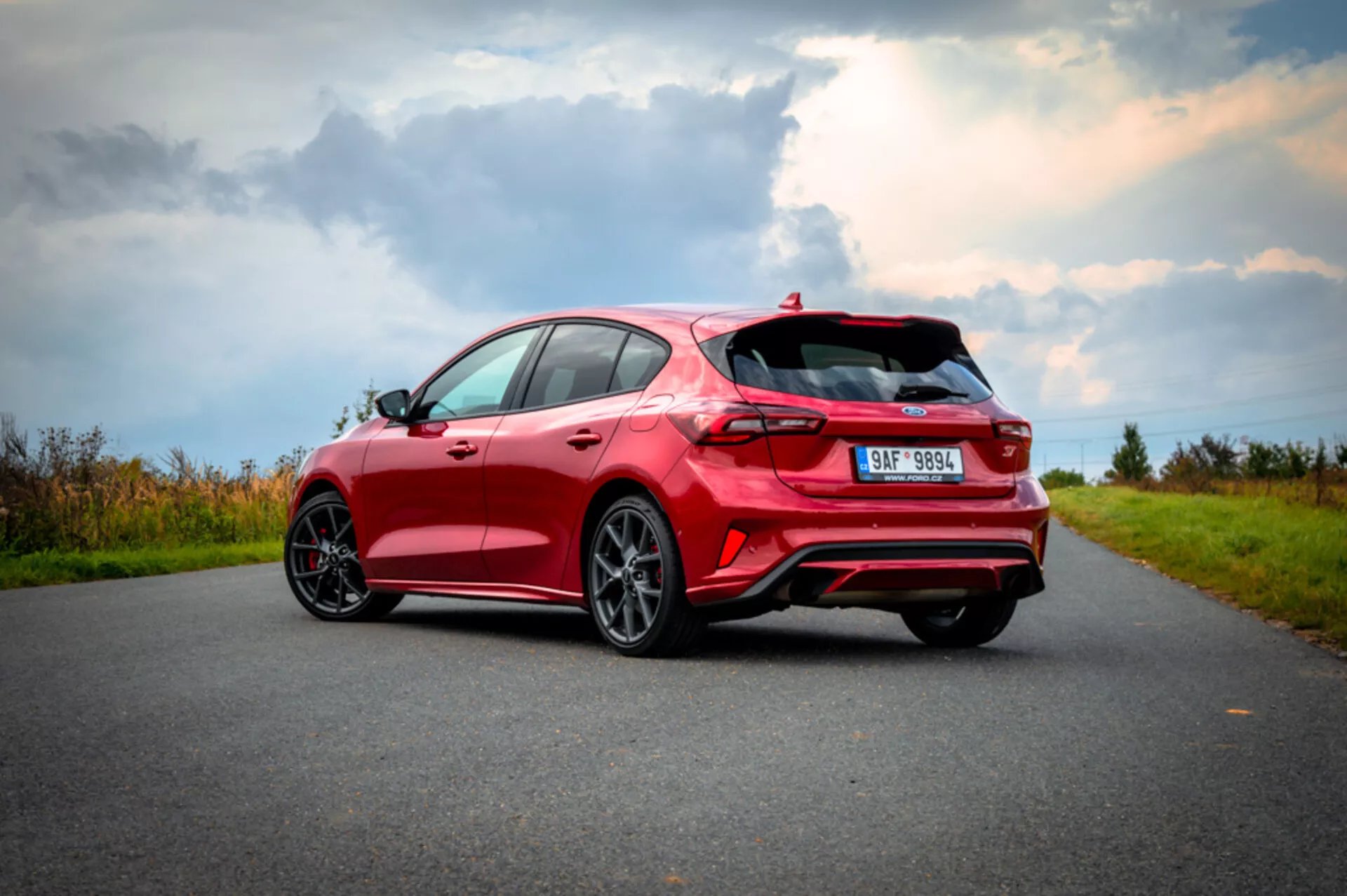 Ford Focus hatchback rosso 2022 visto da angolo posteriore tre quarti che enfatizza il lato destro, con design aerodinamico con spoiler, fanali posteriori LED, doppio scarico, ruote a finitura scura a più razze e pneumatici a basso profilo su strada d'asfalto con paesaggio naturale sullo sfondo sotto cielo parzialmente nuvoloso
