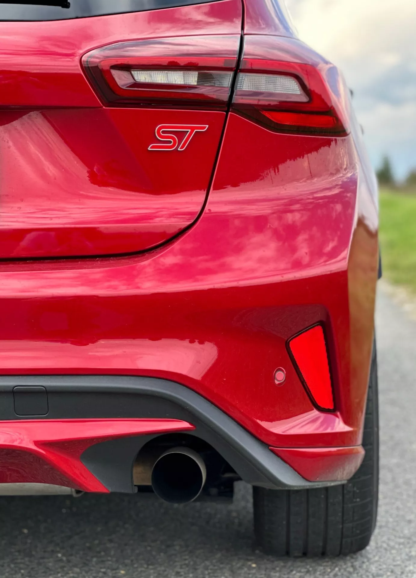 Vista ravvicinata e angolata della sezione posteriore di una Ford Focus 2022 di colore rosso, che mette in evidenza il design e il branding con un emblema ST, una parte del elegante faro posteriore LED e un tubo di scappamento orientato alle prestazioni integrato nel paraurti con un diffusore nero.