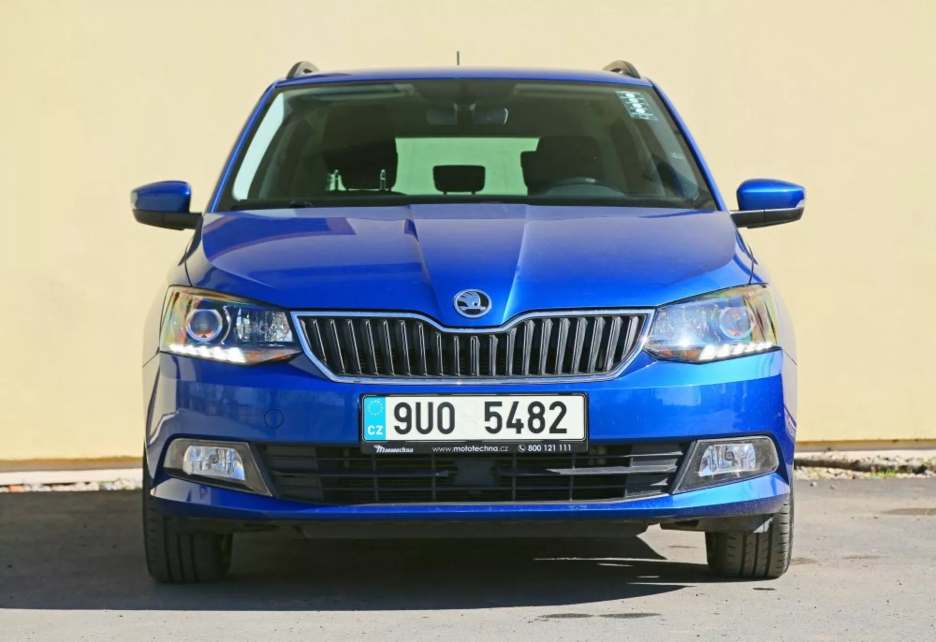 Vista frontale di un veicolo blu Škoda con l'emblema del marchio sulla griglia cromata, fari con lenti trasparenti e fari fendinebbia in alloggiamento nero, con una griglia inferiore dettagliata e un design del cofano scolpito.