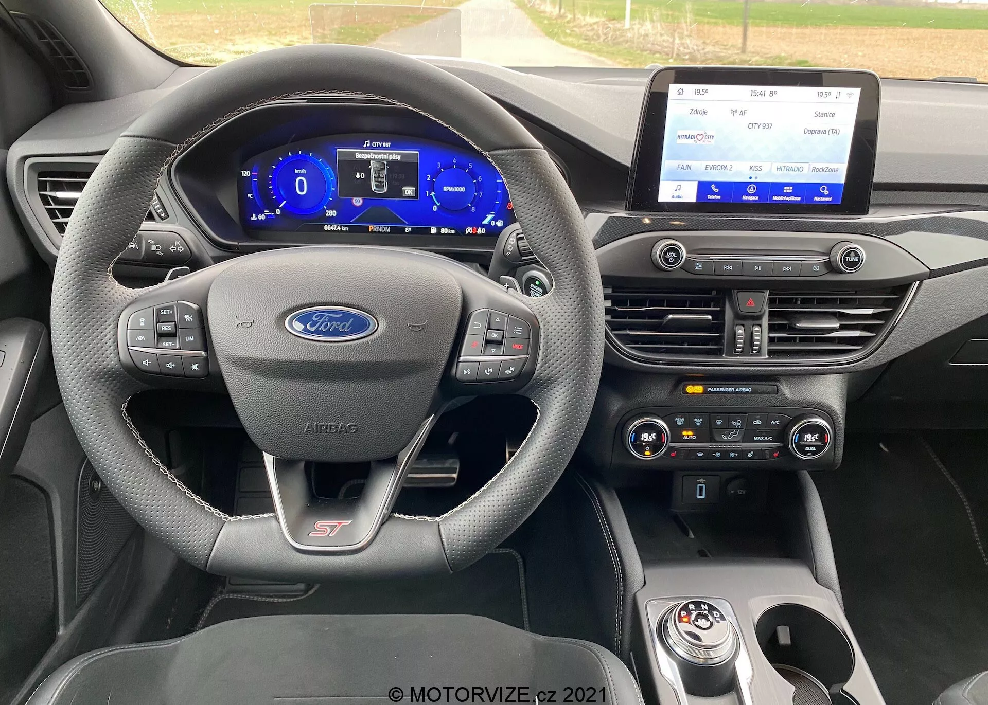 Vista interna di una Ford Focus ST 2022. L'immagine si concentra sul cruscotto dalla prospettiva del conducente. Il volante ha tre razze con comandi multifunzione, rivestito in pelle con cuciture a contrasto. È visibile un badge ST, che indica che è un modello ad alte prestazioni. Dietro al volante, un cruscotto digitale mostra un tachimetro e un contagiri a tema blu. La console centrale è dotata di un grande sistema di infotainment touchscreen orizzontale, controlli del clima a doppia zona e un selettore del cambio rotante con un altro badge ST.