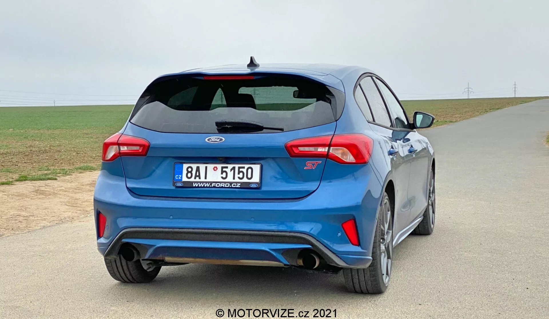 Una Ford Focus 2022 blu hatchback è mostrata da un'angolazione di tre quarti posteriori su una strada asfaltata con un cielo coperto sullo sfondo. L'auto presenta uno spoiler posteriore, luci posteriori moderne, un distintivo sul cofano del bagagliaio con una designazione alfanumerica sotto, un sistema di scarico a doppia uscita, sensori integrati nel paraurti posteriore, un diffusore posteriore, ruote sportive a più raggi, minigonne laterali e un tergicristallo posteriore. L'immagine mette in evidenza il design e le caratteristiche di prestazione dell'auto in un possibile contesto rurale.