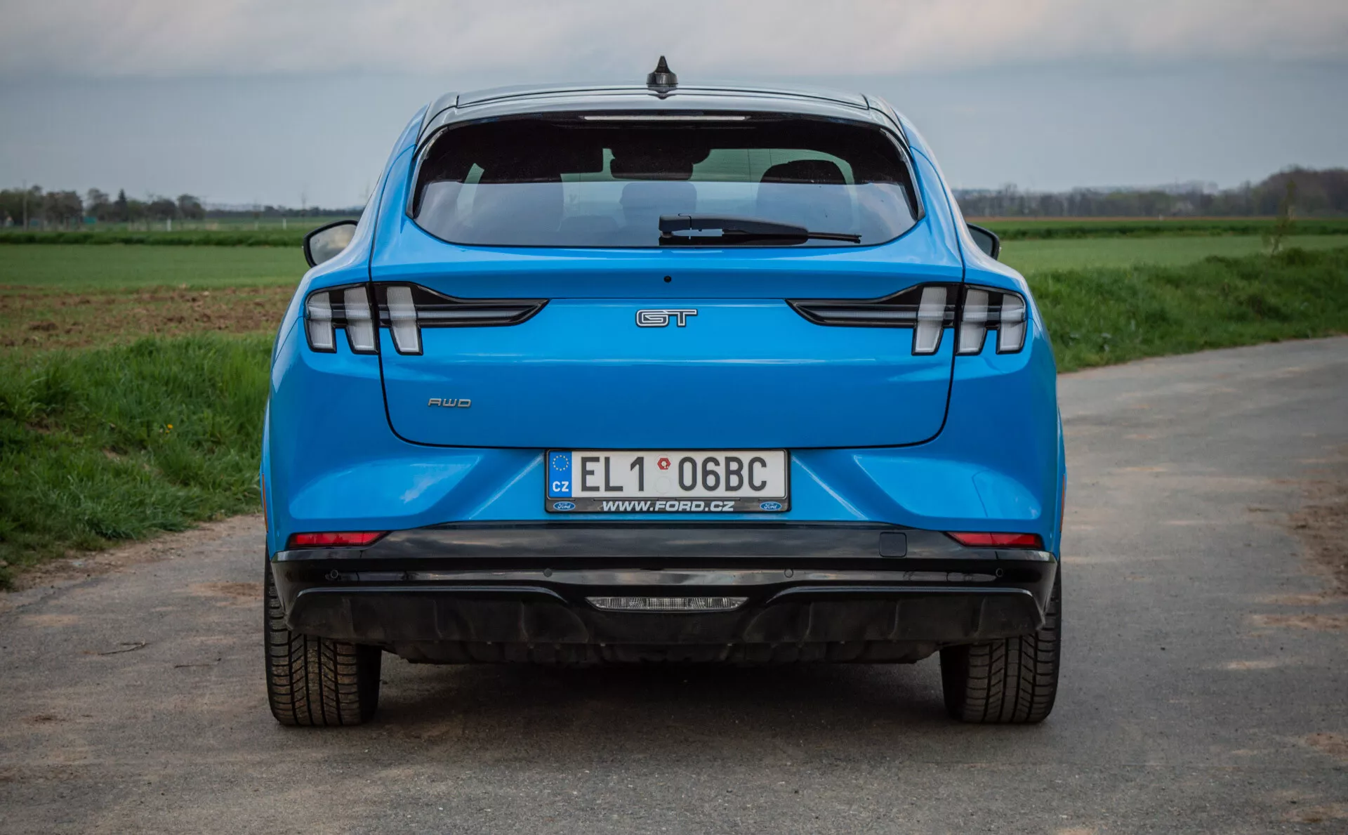 Vista posteriore di una Ford Mustang-Mach-E blu del 2020 su una strada asfaltata con campi d'erba sullo sfondo, che mostra caratteristiche chiave come il logo del produttore, il nome del modello, il badge del livello di allestimento, la targa polacca, sistema di scarico doppio, fanali posteriori LED moderni, sensori di parcheggio e una luce di stop alta montata.