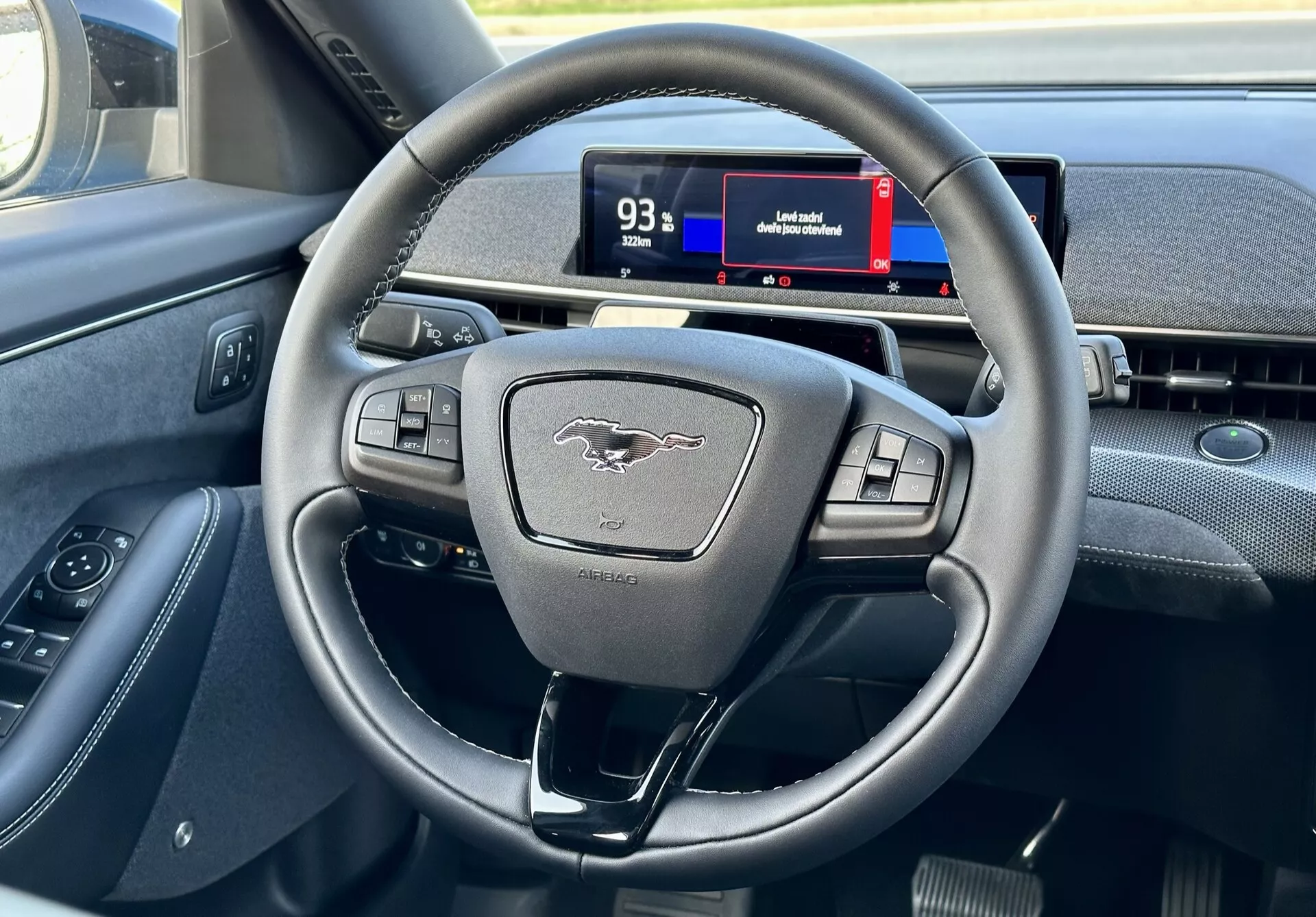 Interno della Ford Mustang Mach-E 2020 che evidenzia il volante moderno a tre razze con pulsanti multifunzione e il quadro strumenti digitale dalla prospettiva del conducente, mostrando comandi, emblema e accenni di navigazione.