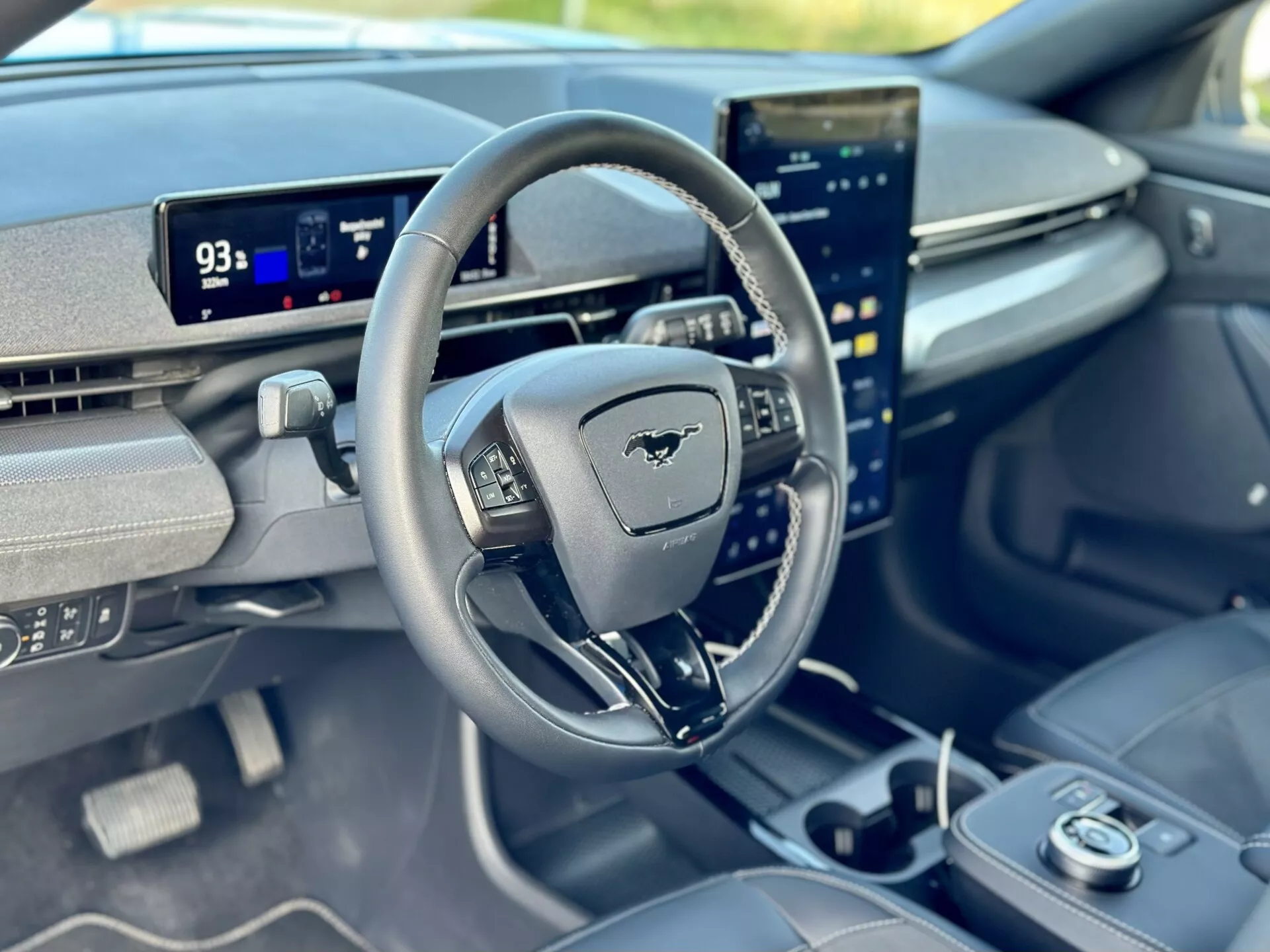 Vista interna di una Ford Mustang Mach-E 2020 concentrata sul cockpit del guidatore dalla prospettiva del passeggero anteriore, presentando un volante con emblema Mustang, cruscotto digitale in modalità Parcheggio con autonomia di 93 miglia, e un touchscreen centrale orientato verticalmente integrato con capacità avanzate di infotainment.