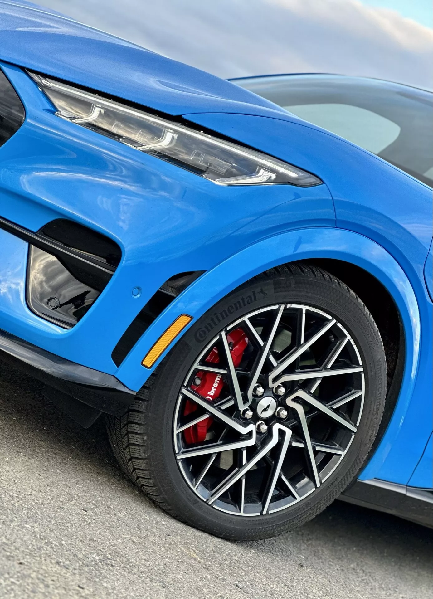Primo piano dettagliato del Ford Mustang Mach-E 2020 che mette in mostra il lato anteriore sinistro, concentrato sulla ruota in lega con pinza freno rossa, parafango anteriore slanciato con una presa d'aria di raffreddamento, vista parziale del moderno faro LED, colore blu sportivo della carrozzeria e miglioramenti al trim esterno
