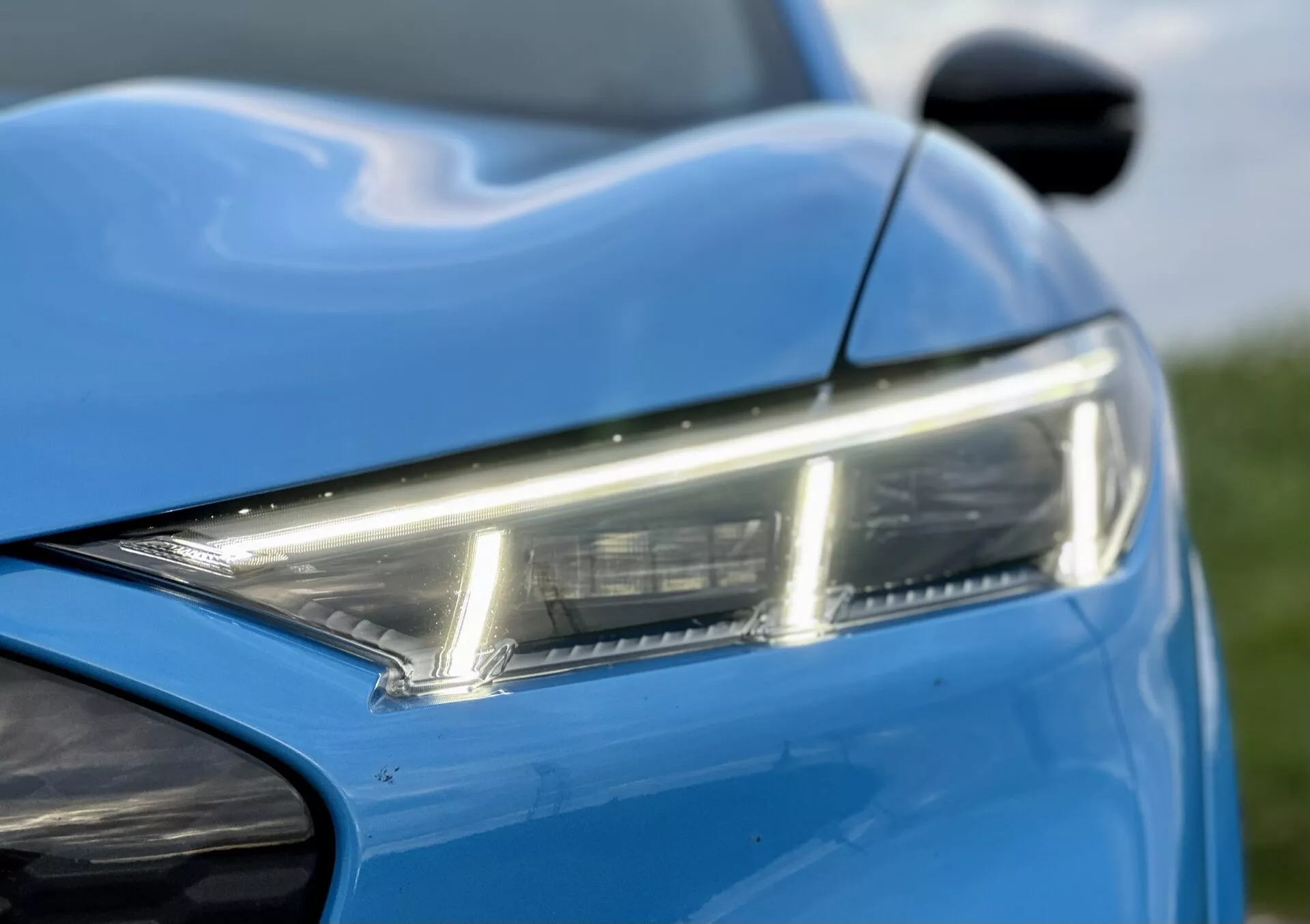 Vista ravvicinata della sezione anteriore sinistra del Ford Mustang-Mach-E 2020, evidenziando il faro LED elegante, la vernice blu vibrante e le linee dinamiche della carrozzeria, con uno scorcio dello specchietto laterale e del paraurti anteriore, per mostrare il design moderno e l'estetica.