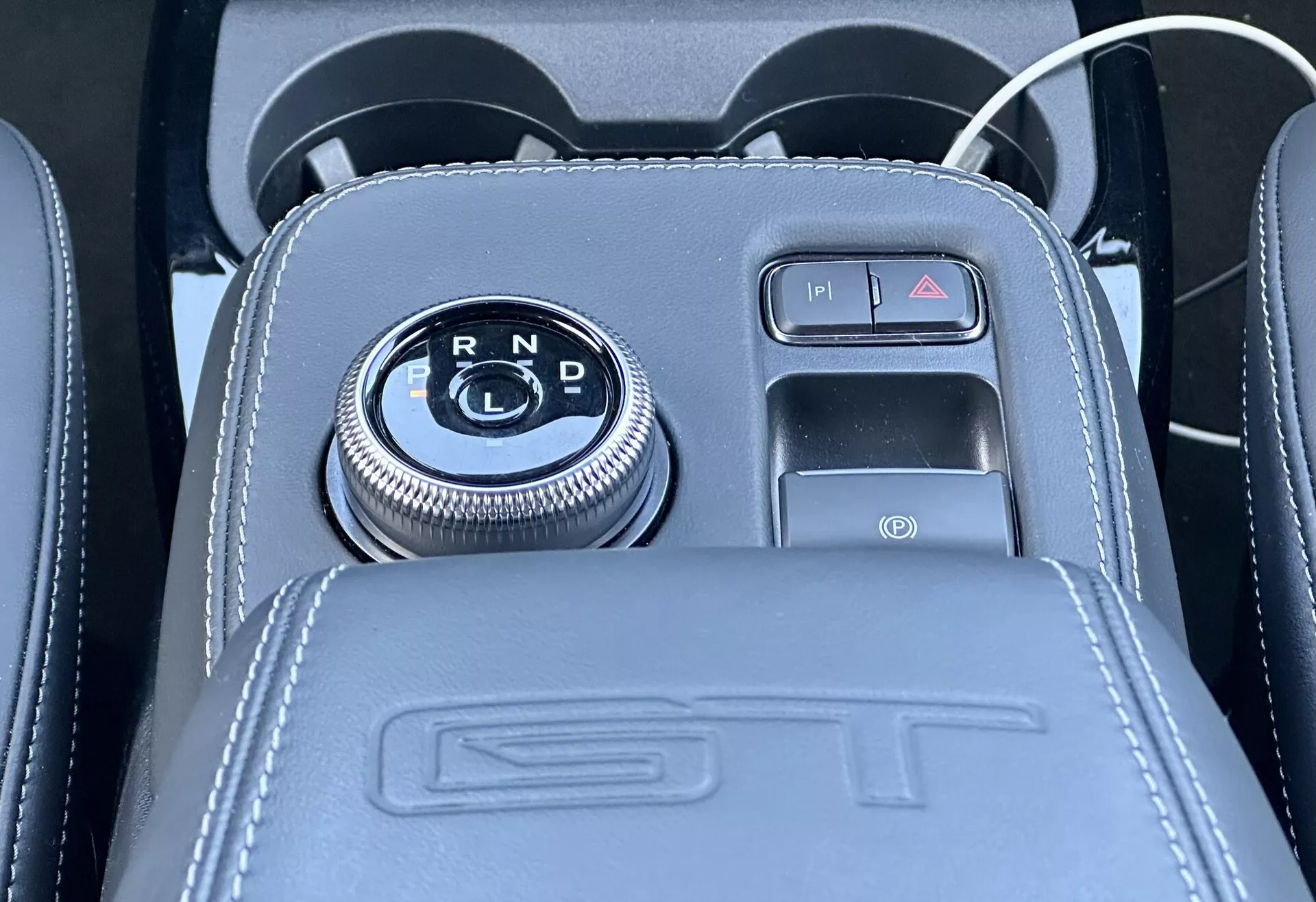 Vista ravvicinata della console centrale della Ford Mustang Mach-E 2020 con selettore rotativo del cambio posizionato su un pannello nero lucido con indicatori di marcia R, N, D, circondato da una finitura cromata, accanto a un freno di parcheggio elettronico con pulsante con dettagli in pelle, goffratura ST e inclusi una porta USB e una presa per accessori 12V
