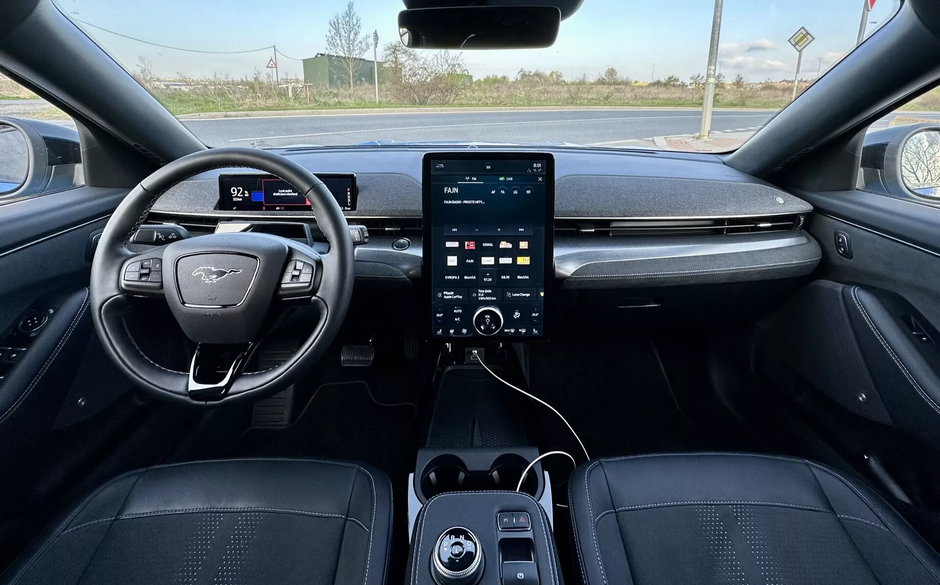 Vista in prima persona dell'interno della Ford Mustang Mach-E 2020, mostrando il volante posizionato a sinistra con design a fondo piatto e emblema, cluster strumenti digitale, grande sistema di infotainment con touchscreen verticale sulla console centrale con sotto i pulsanti di controllo clima, porte USB e rivestimento di lusso in toni scuri con cuciture dettagliate, suggerendo caratteristiche di sicurezza e assistenza avanzate, con uno sfondo esterno rurale visibile attraverso il parabrezza.