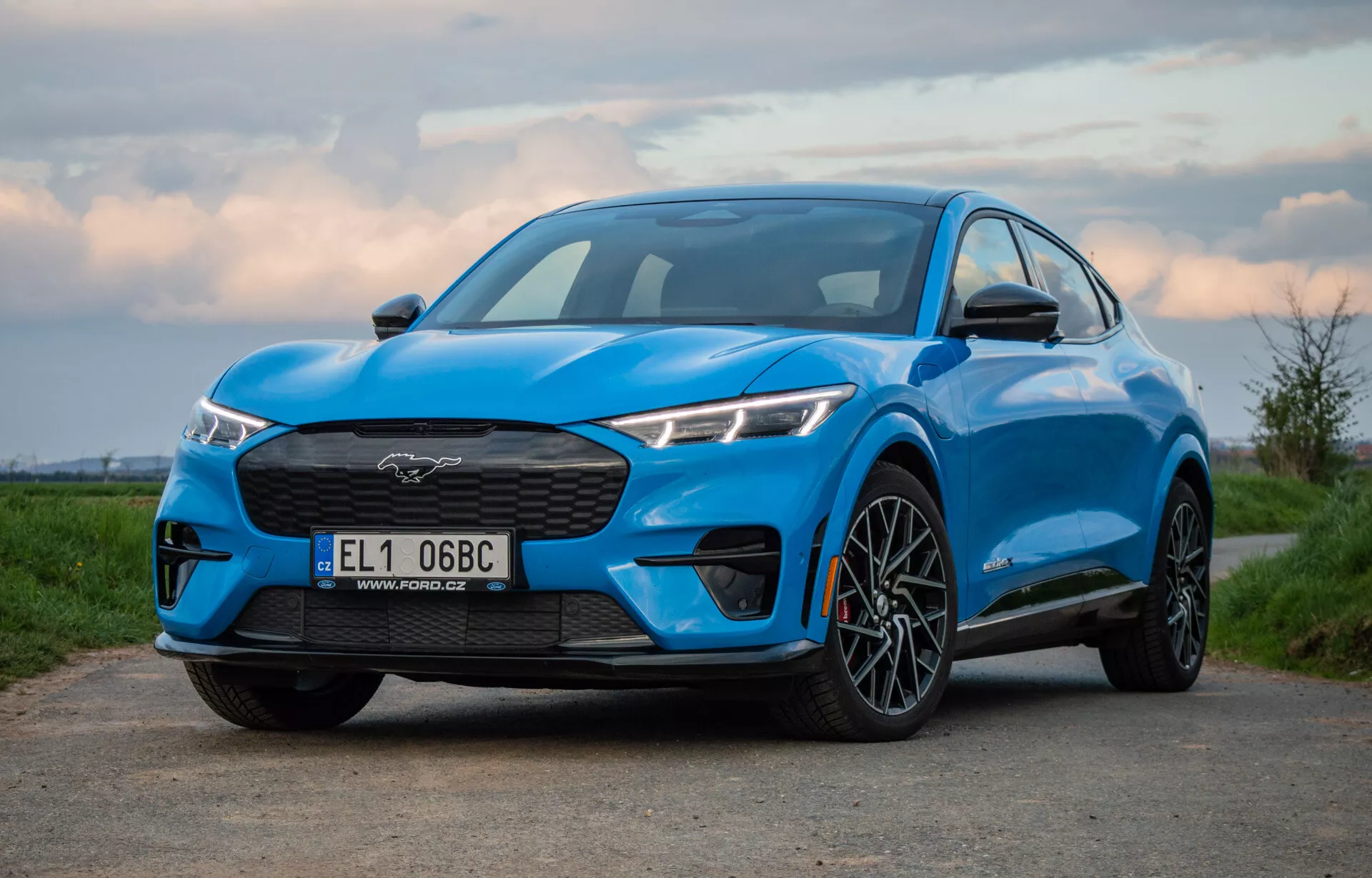 Ford Mustang Mach-E blu 2020 fotografato da un angolo basso e leggermente decentrato che mostra la griglia, i fari, il cofano e il profilo del SUV elettrico incluso la ruota anteriore e il parafango, sottolineando il suo design aerodinamico e performante all'alba o al tramonto.