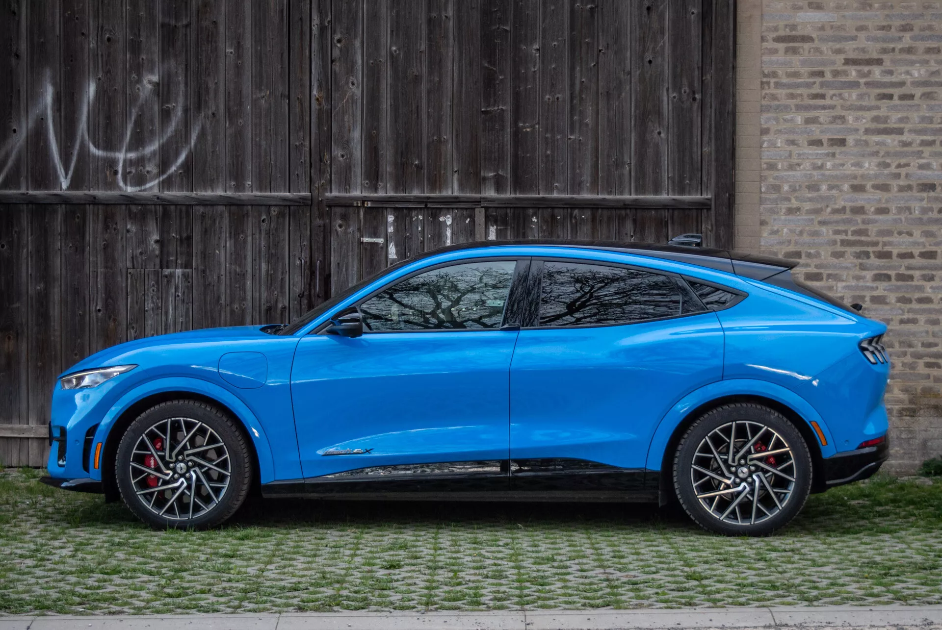 SUV crossover blu Ford Mustang Mach-E 2020 parcheggiato parallelamente a un cancello di garage rustico in legno con graffiti, vista laterale che mostra il design aerodinamico, tetto inclinato in stile coupé, modello a quattro porte con maniglie integrate, specchietti laterali aerodinamici con segnali di svolta, grandi ruote in lega bicolore con pinze dei freni rosse, altezza da terra moderata, carrozzeria sagomata con linee scolpite e passaruota pronunciati, linea dei finestrini affusolata con piccola finestra dopo il pilastro C e minigonne laterali sportive