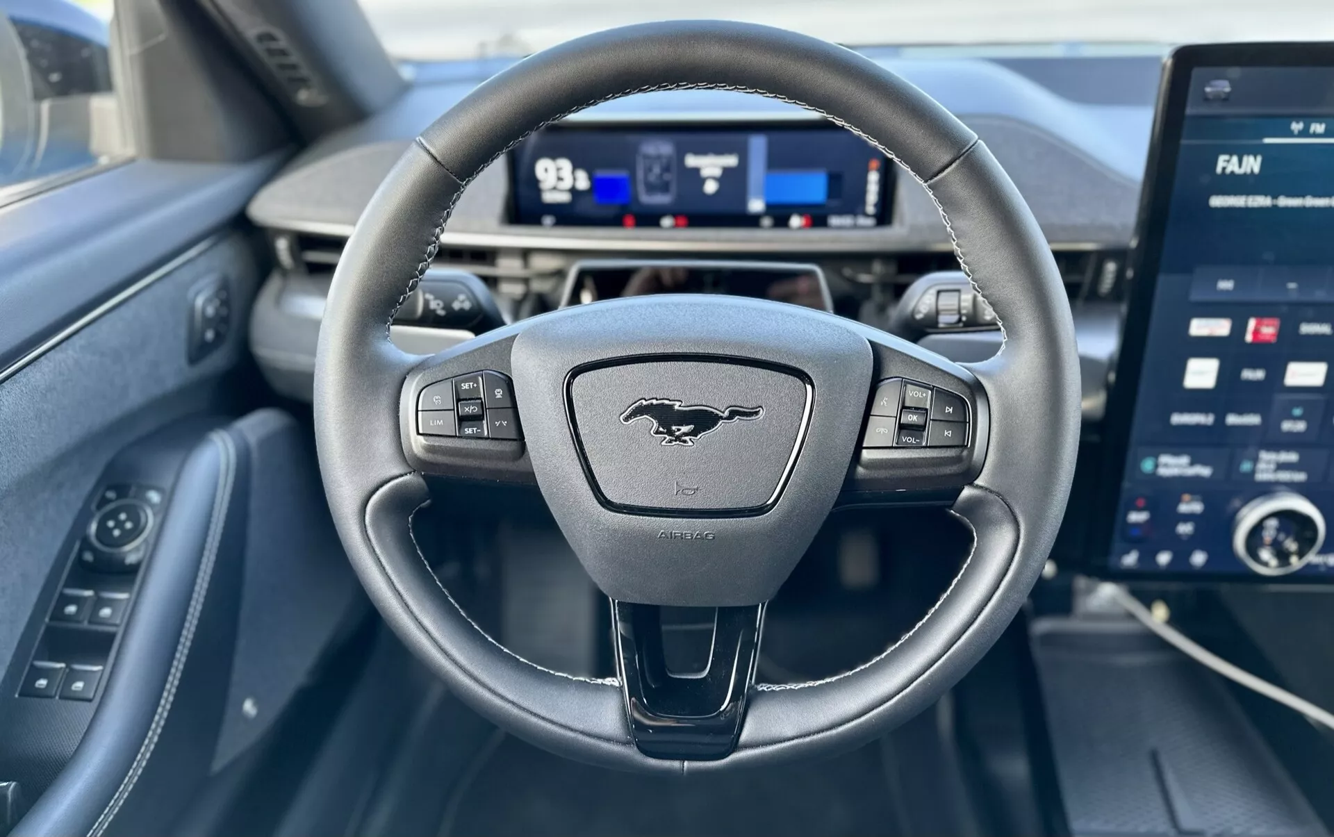Vista interna di una Ford Mustang-Mach-E 2020 dalla prospettiva del conducente, caratterizzata da un volante a tre razze con comandi, un cruscotto digitale e un ampio touchscreen per l'infotainment