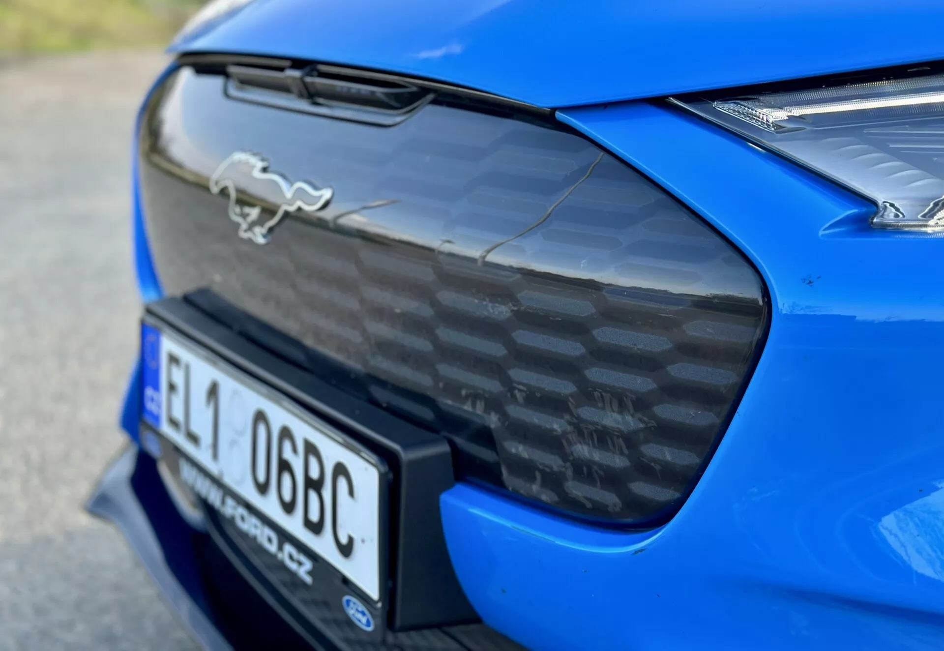 Primo piano della sezione posteriore di una Ford Mustang Mach-E 2020 blu, con emblema del cavallo cromato, spoiler posteriore in fibra di carbonio, nicchia integrata per la targa e visuale parziale delle luci posteriori LED, che evidenziano le caratteristiche aerodinamiche e di prestazione dell'auto.