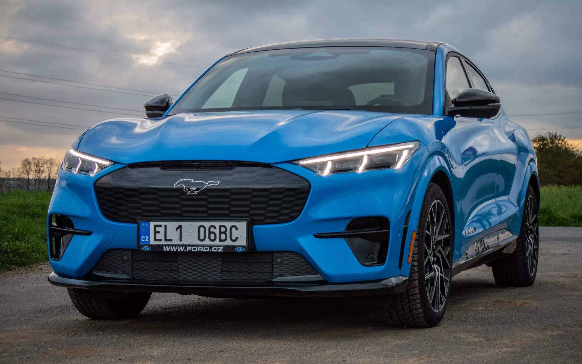 Ford Mustang Mach-E 2020 in blu brillante che mostra un design sportivo e aerodinamico da una prospettiva frontale e laterale bassa, evidenziando fari LED angolari, griglia chiusa, cofano sagomato e ruote in lega a più razze contro uno sfondo naturale.