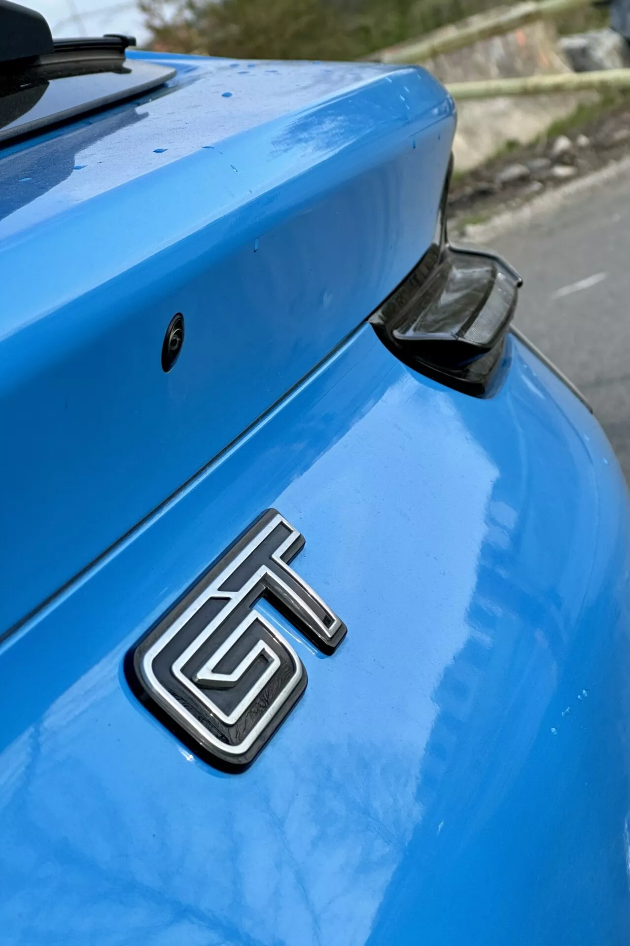 Primo piano della sezione posteriore di una Ford Mustang Mach-E blu del 2020 con vista dettagliata del distintivo GT metallico, del bagagliaio curvo e dello spoiler posteriore aerodinamico con tappo finale nero, su uno sfondo delicatamente sfocato.