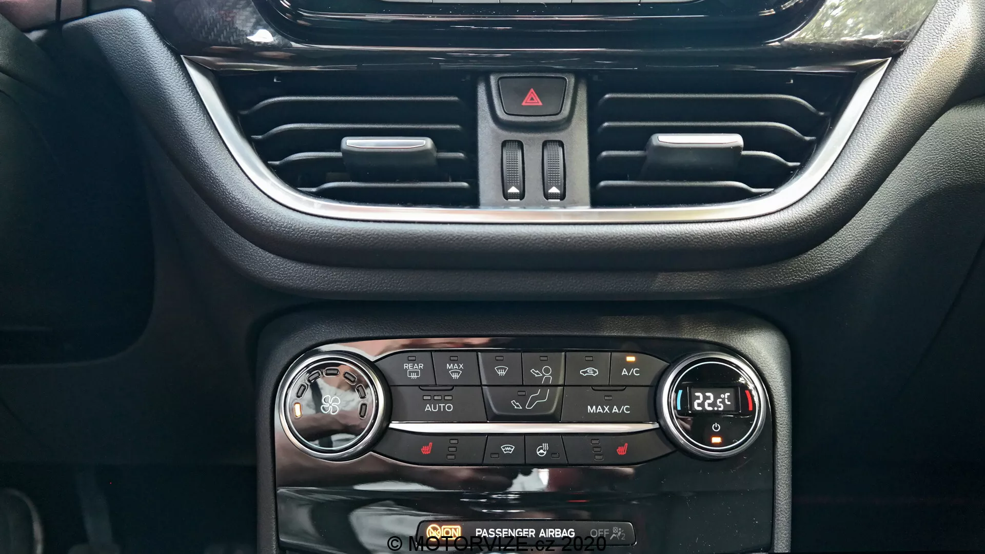 L'immagine cattura la console centrale del cruscotto della Ford Puma 2020, evidenziando il sistema di controllo del clima e parte del sistema di infotainment. Da un angolo frontale, la bocchetta d'aria centrale con alette orizzontali regolabili è visibile nella parte superiore. Sotto c'è il pulsante triangolare rosso delle luci di emergenza. Il pannello di controllo del clima mostra una varietà di pulsanti e due manopole per impostare la temperatura e il flusso d'aria, con funzioni di controllo del clima AUTO, A/C, controllo a doppia zona, sbrinamento posteriore e ricircolo. Ogni manopola della temperatura ha un display digitale, impostato su 22,5 gradi Celsius. Sul bordo inferiore è presente l'indicatore 'AIRBAG PASSEGGERO DISATTIVATO'. Il design interno integra plastiche nere moderne e accenti argentati per un'interfaccia intuitiva.