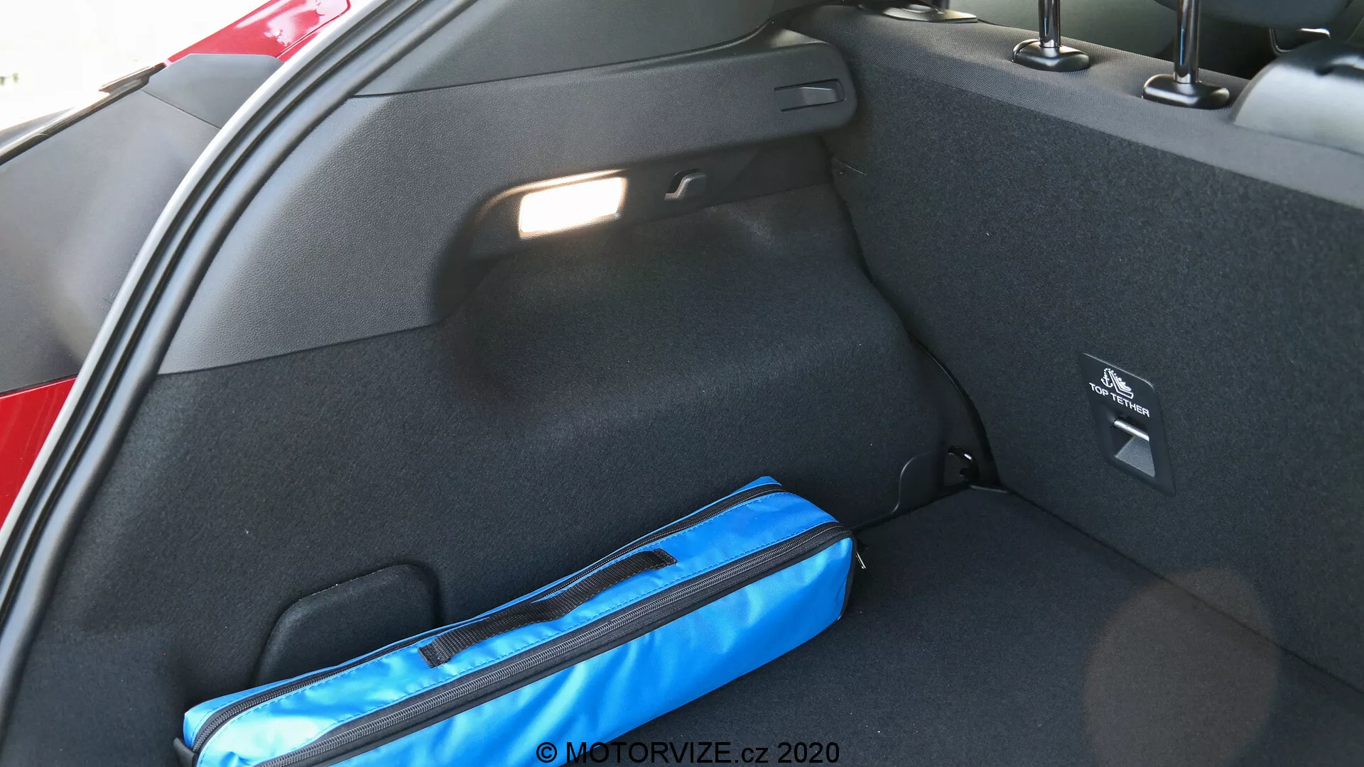 Vista ravvicinata dell'interno del bagagliaio di un'auto dal lato, concentrandosi sulla parte destra. L'area del carico è rifinita in materiale di moquette nera e presenta un vano portaoggetti laterale con un'etichetta di sicurezza. Un oggetto blu, possibilmente una borsa o un organizzatore, si trova sul pavimento. La luce naturale illumina il bagagliaio, indicando che è aperto.