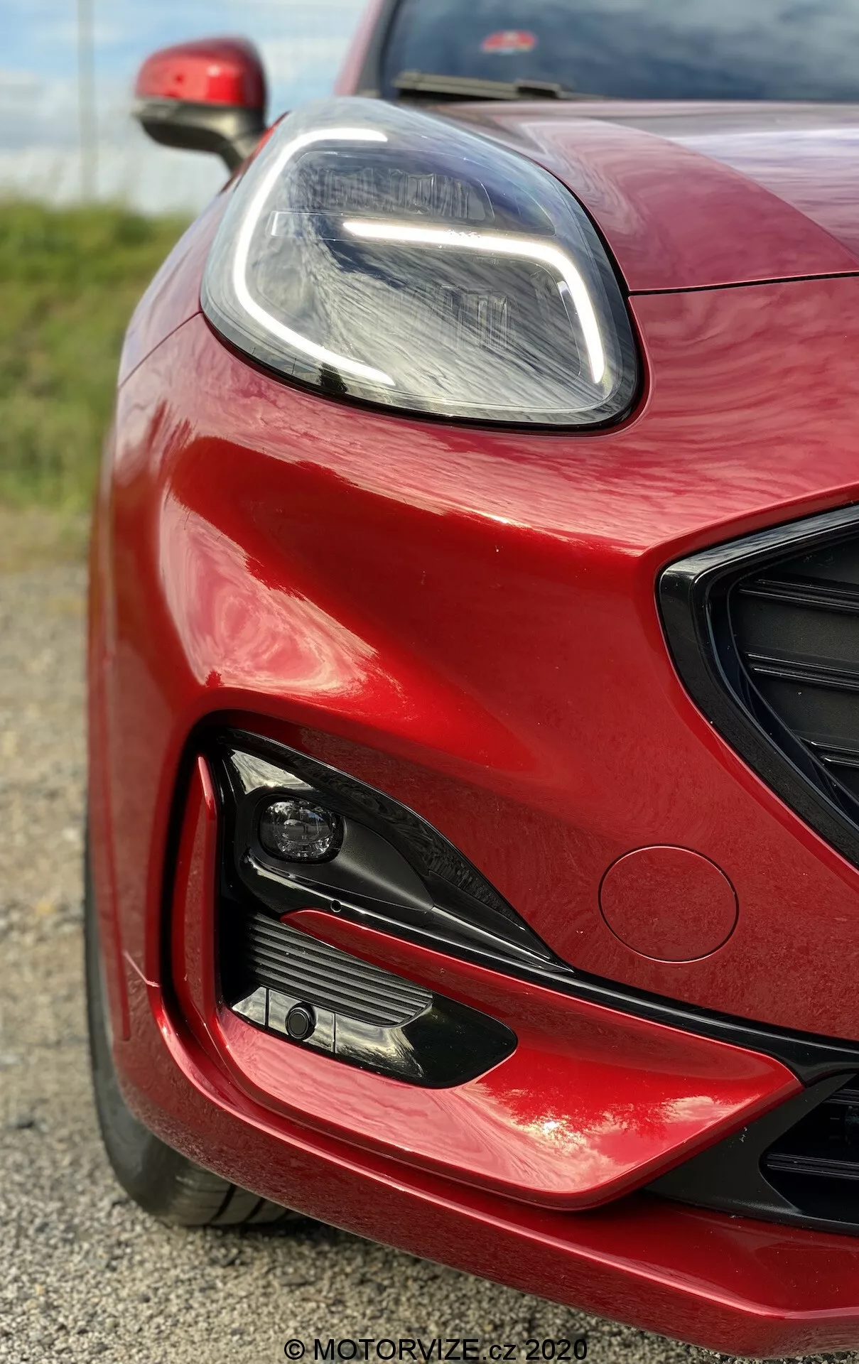 Primo piano della parte anteriore sinistra di una Ford Puma 2020 in rosso vivace con finitura lucida, che mostra il elegante faro a LED, grandi prese d'aria e fendinebbia, enfatizzando il suo design moderno e orientato alle prestazioni.