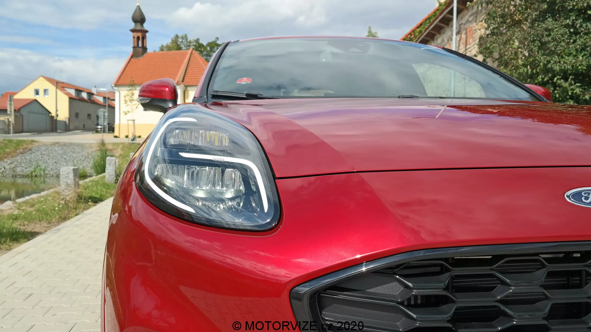 Vista frontale ravvicinata di una Ford Puma rossa 2020, che mostra il faro destro con elementi LED eleganti, il cofano scolpito aerodinamico e la griglia a nido d'ape. Immagine scattata da un'angolazione bassa, che cattura lo stile moderno e i dettagli sportivi dell'auto.