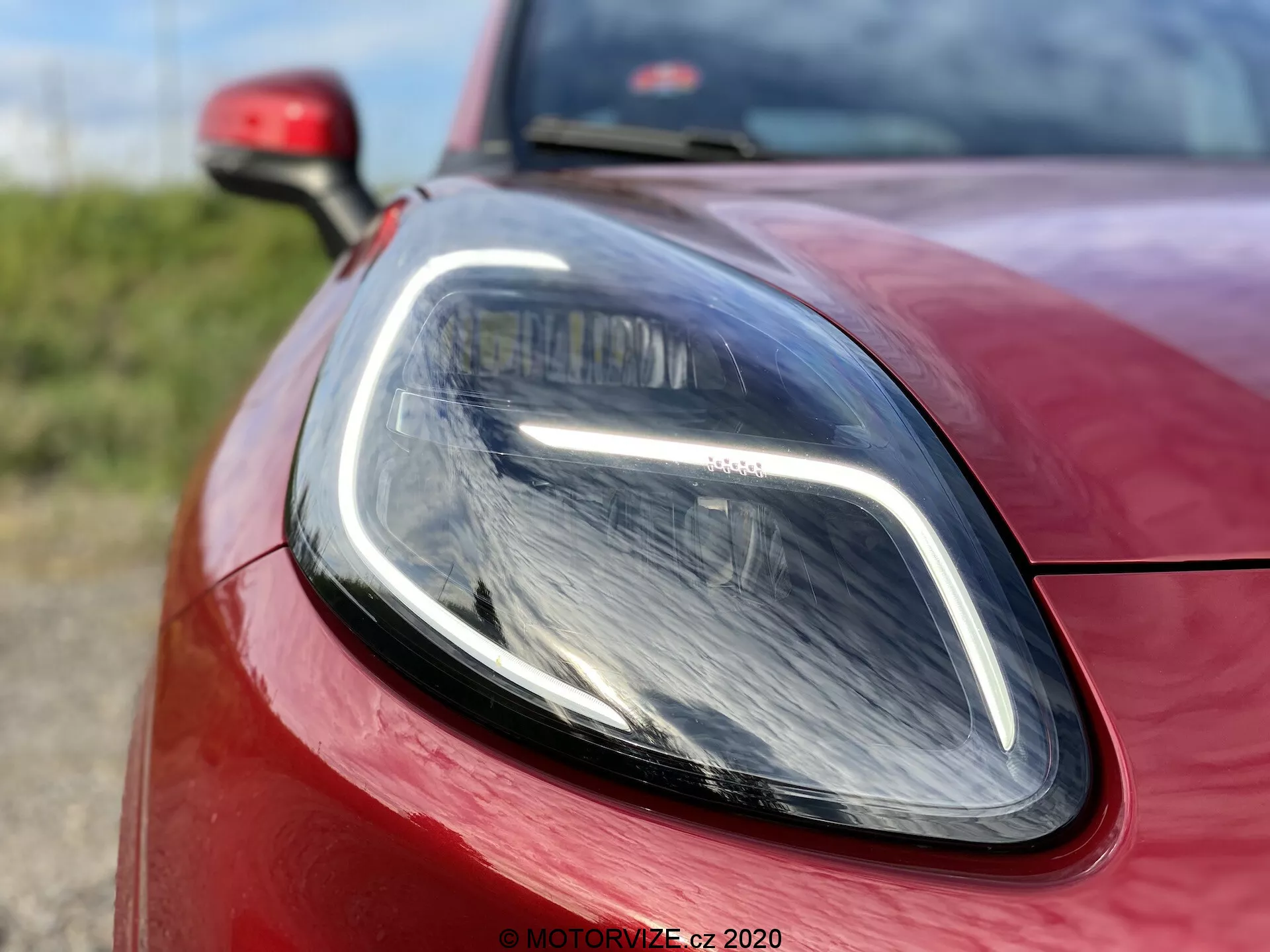 Immagine ravvicinata della Ford Puma 2020, focalizzata sulla sezione del faro anteriore lato conducente e sulla carrozzeria circostante. Scattata da un angolo basso e leggermente decentrato, l'immagine mette in risalto le luci diurne LED eleganti e il faro di tipo proiettore moderno. La vernice rossa vibrante e l'allineamento preciso dei pannelli enfatizzano il design sportivo e aerodinamico dell'auto.