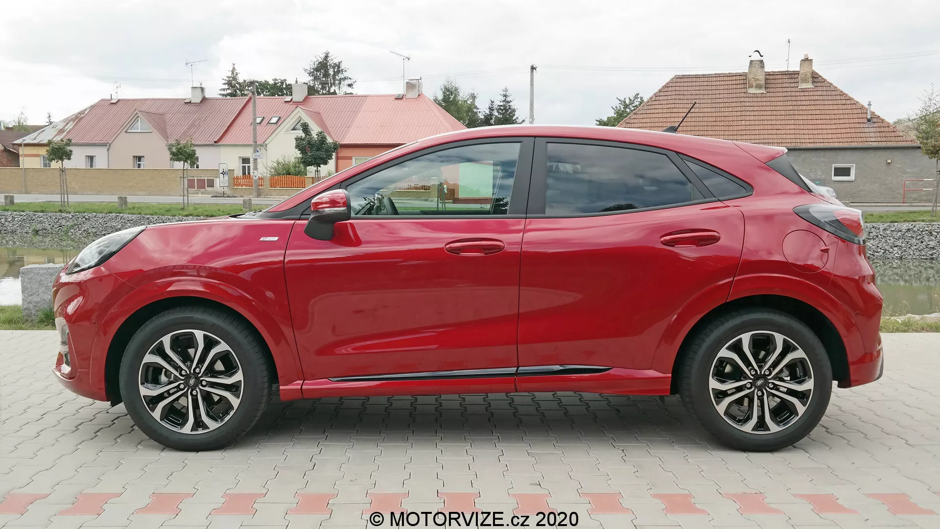 Vista laterale di profilo di una Ford Puma hatchback rossa a quattro porte del 2020, scattata durante il giorno in un parcheggio con uno sfondo di erba ed edifici residenziali. L'auto presenta linee aerodinamiche, cerchi in lega a più razze, specchietti laterali e maniglie delle porte in tinta, e una linea del tetto discendente. L'assenza di serrature visibili suggerisce un possibile sistema di accesso senza chiave.