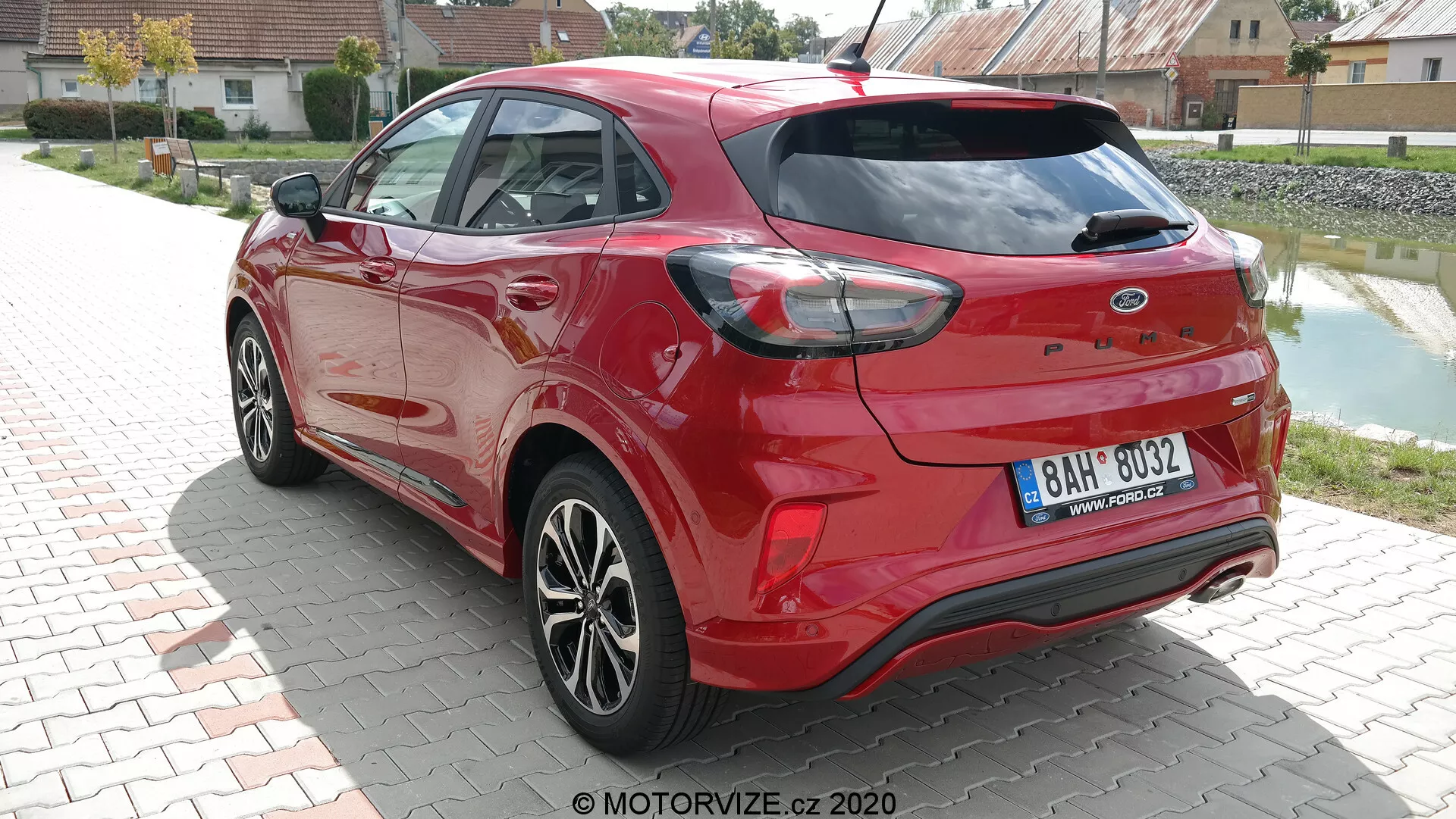 Vista posteriore di tre quarti di una Ford Puma 2020 hatchback di colore rosso vivace catturata alla luce del giorno. L'immagine mette in risalto i contorni aerodinamici dell'auto, le caratteristiche luci posteriori che si avvolgono intorno ai lati e i sensori integrati nel paraurti posteriore, suggerendo capacità di assistenza al parcheggio. Le doppie punte di scarico e lo spoiler posteriore aumentano il suo fascino sportivo, mentre le ruote in lega multirazze e gli elementi colorati della carrozzeria, come gli specchietti laterali e le maniglie delle porte, aggiungono al design moderno. Spazzola tergicristallo posteriore visibile, indicando praticità in condizioni meteorologiche avverse.