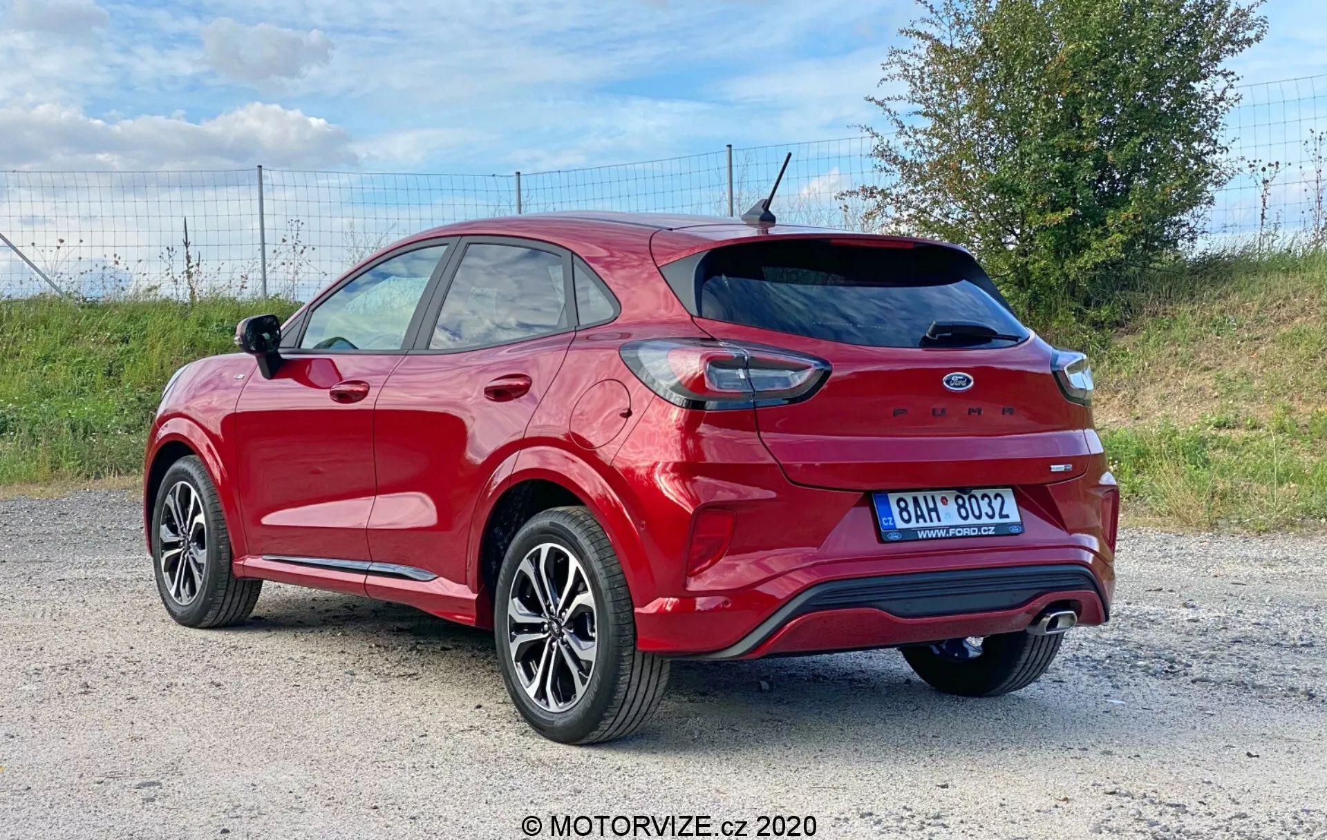 L'immagine presenta una Ford Puma 2020 crossover/SUV rossa fotografata da un angolo di tre quarti posteriore, mostrando il pneumatico posteriore e la ruota, parte della porta anteriore e della porta passeggeri posteriore, il pannello posteriore e tutto il portellone con spoiler integrato e tergicristallo posteriore. Le caratteristiche visibili includono luci posteriori parzialmente avvolgenti intorno al parafango posteriore, paraurti posteriore con riflettori e sensori di parcheggio, sistema di scarico doppio, antenna a pinna di squalo, parabrezza posteriore con linee di sbrinamento, ruote in lega con design a più razze e pneumatici a basso profilo, vetri oscurati, barre portatutto sul tetto e calotta dello specchietto laterale in tinta con la carrozzeria. Il veicolo è situato in un ambiente esterno, probabilmente su una strada sterrata o di ghiaia, con un tratto di prato visibile sullo sfondo che suggerisce un'ambientazione rurale o semi-rurale.