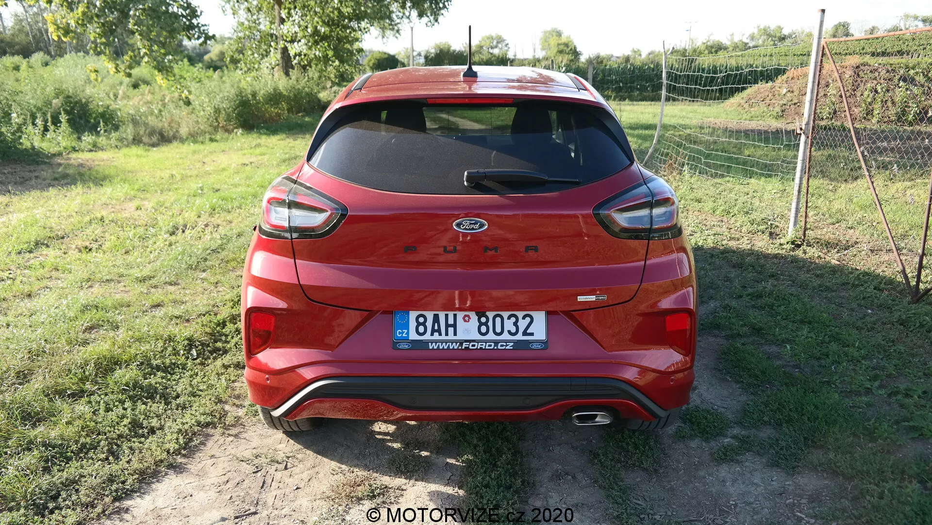 Vista posteriore di una Ford Escape rossa del 2020 con tecnologia ibrida parcheggiata su una superficie non asfaltata. Il veicolo presenta luci posteriori moderne, logo Ford centrato e targa europea. Il parabrezza posteriore ha un tergicristallo e una terza luce di stop a LED. La parte inferiore della carrozzeria e il paraurti hanno rifiniture protettive in plastica nera con sensori visibili, che indicano la tecnologia di assistenza al parcheggio. La vegetazione sullo sfondo suggerisce un ambiente rurale.