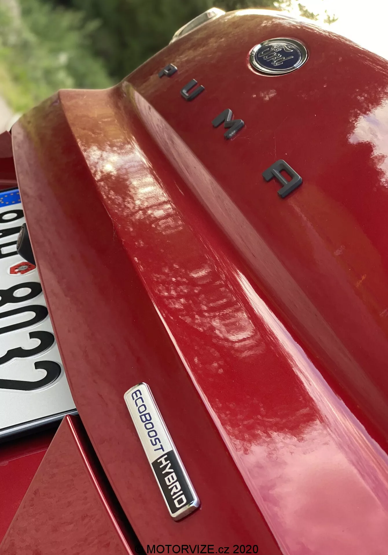Dettaglio ravvicinato del quarto posteriore sinistro di una Ford Puma 2020 rossa, ripreso da un'angolazione verso l'alto, mettendo in evidenza distintivi e marchi. L'immagine mette in risalto l'emblema della Ford in alto, il nome del modello 'PUMA' in lettere cromate sul baule e il distintivo 'ECOBOOST HYBRID', che indica una tecnologia ibrida avanzata. Una parte della targa è parzialmente visibile in basso a sinistra. La vernice lucida riflette la luce, accentuando il design e la sofisticatezza tecnologica del veicolo.