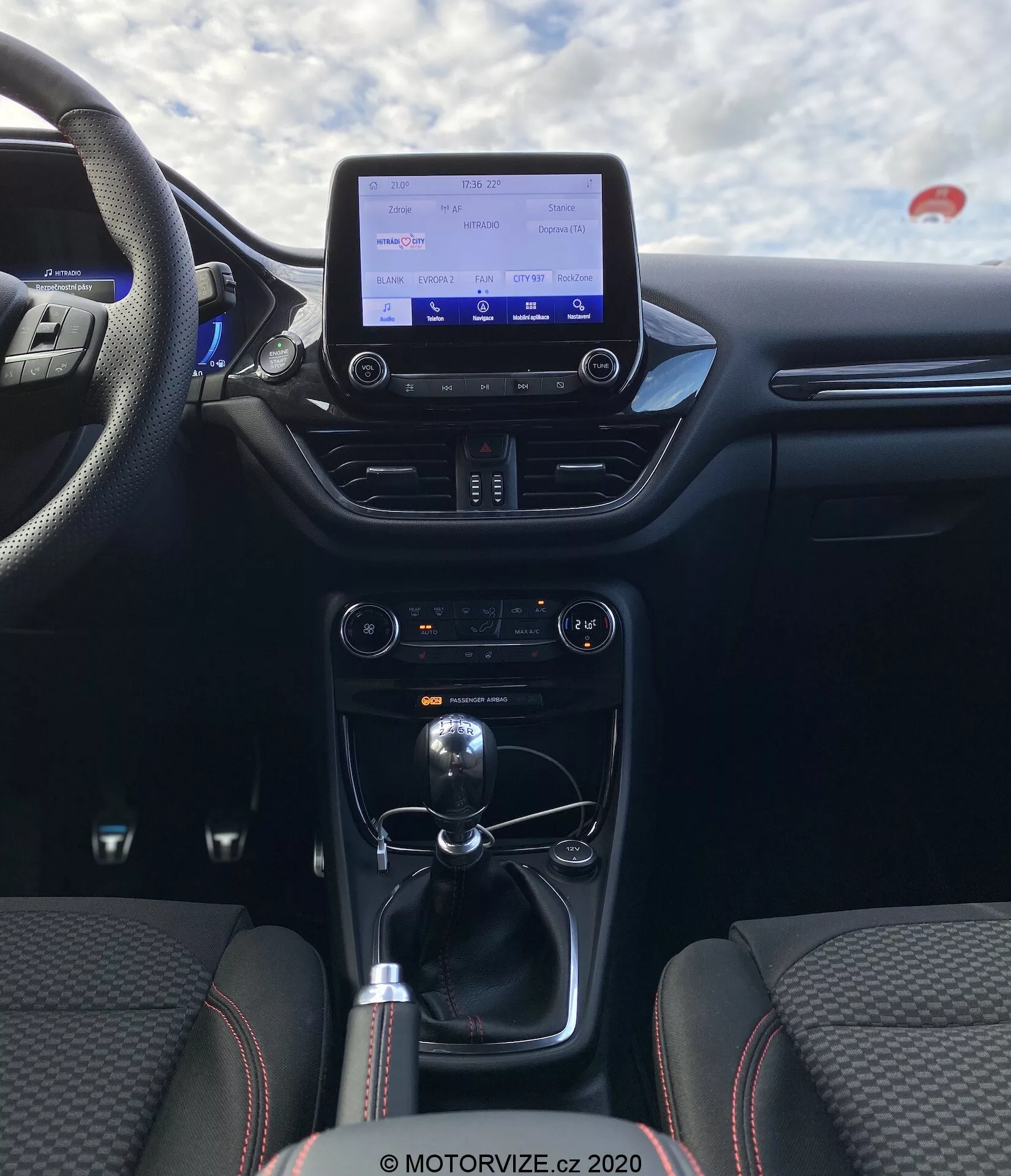 Vista dell'interno della Ford Puma 2020 dalla prospettiva del passeggero posteriore, concentrandosi sull'area della cabina centrale. L'immagine mette in risalto la console centrale e il cruscotto, includendo un display di infotainment, sistema di controllo del clima, leva del cambio per trasmissione manuale e opzioni di connettività come porte USB e presa da 12V. I sedili sono rivestiti in tessuto con un motivo di cuciture distintivo e con rinforzi laterali, enfatizzando comfort e sportività.