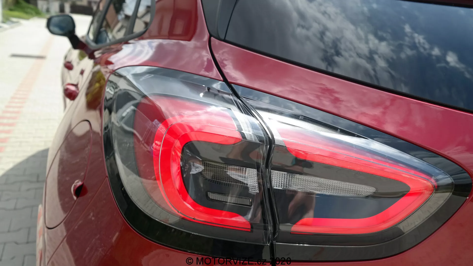 Primo piano della sezione posteriore di una Ford Puma 2020, con messa a fuoco sul moderno design delle luci posteriori a LED da un angolo leggermente elevato. La luce del freno rossa, i segmenti chiari e scuri per la retromarcia e gli indicatori di direzione si fondono perfettamente con la vernice rosso scuro o bordeaux ad alta lucidità dell'auto, riflettendo l'ambiente circostante. L'immagine mette in evidenza il design curvilineo e contemporaneo dell'auto e la sua tecnologia di illuminazione funzionale.