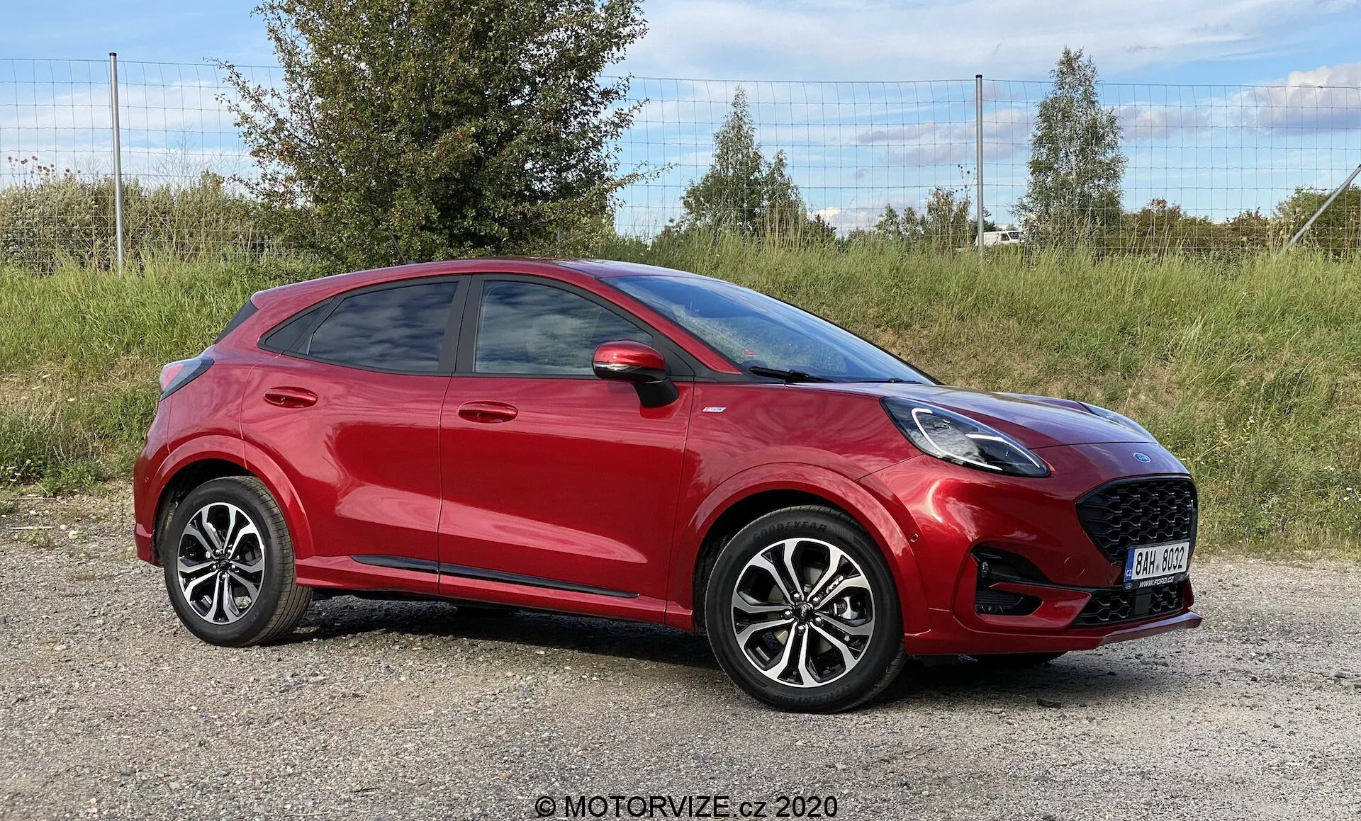Fotografia professionale di un SUV crossover compatto Ford Puma 2020 rosso, posizionato su un terreno erboso con fogliame sullo sfondo. L'angolo anteriore laterale mette in risalto la prominente griglia anteriore a nido d'ape, i fari LED eleganti e lo specchietto laterale parzialmente visibile. La linea del tetto inclinata sportiva, le linee distintive delle porte, le maniglie chiaramente visibili, i cerchi in lega con design a più raggi e gli pneumatici a profilo basso contribuiscono all'estetica moderna dell'auto. L'immagine, scattata a livello dell'auto, cattura tutta la sua lunghezza, enfatizzando il design contemporaneo.