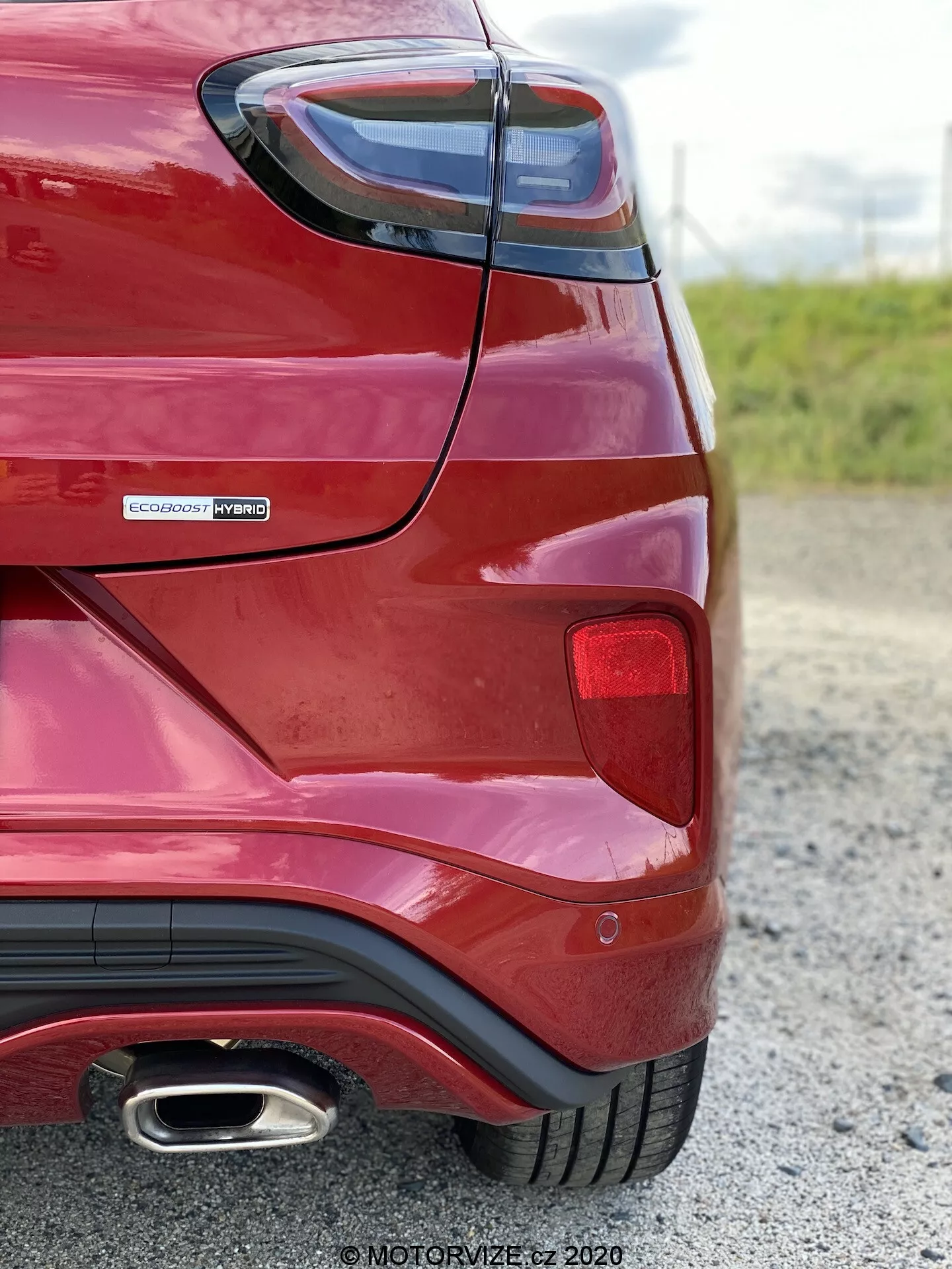 Vista angolare posteriore di una Ford Puma 2020 rossa scattata da vicino e da bassa angolazione, con particolare attenzione alla curvatura del parafango posteriore, il gruppo luci posteriori, il badge EcoBoost Hybrid che indica la motorizzazione ibrida e il singolo terminale di scarico cromato. Evidenziando elementi di design moderni come luci posteriori angolari e paraurti posteriore aerodinamico con stile diffusore.