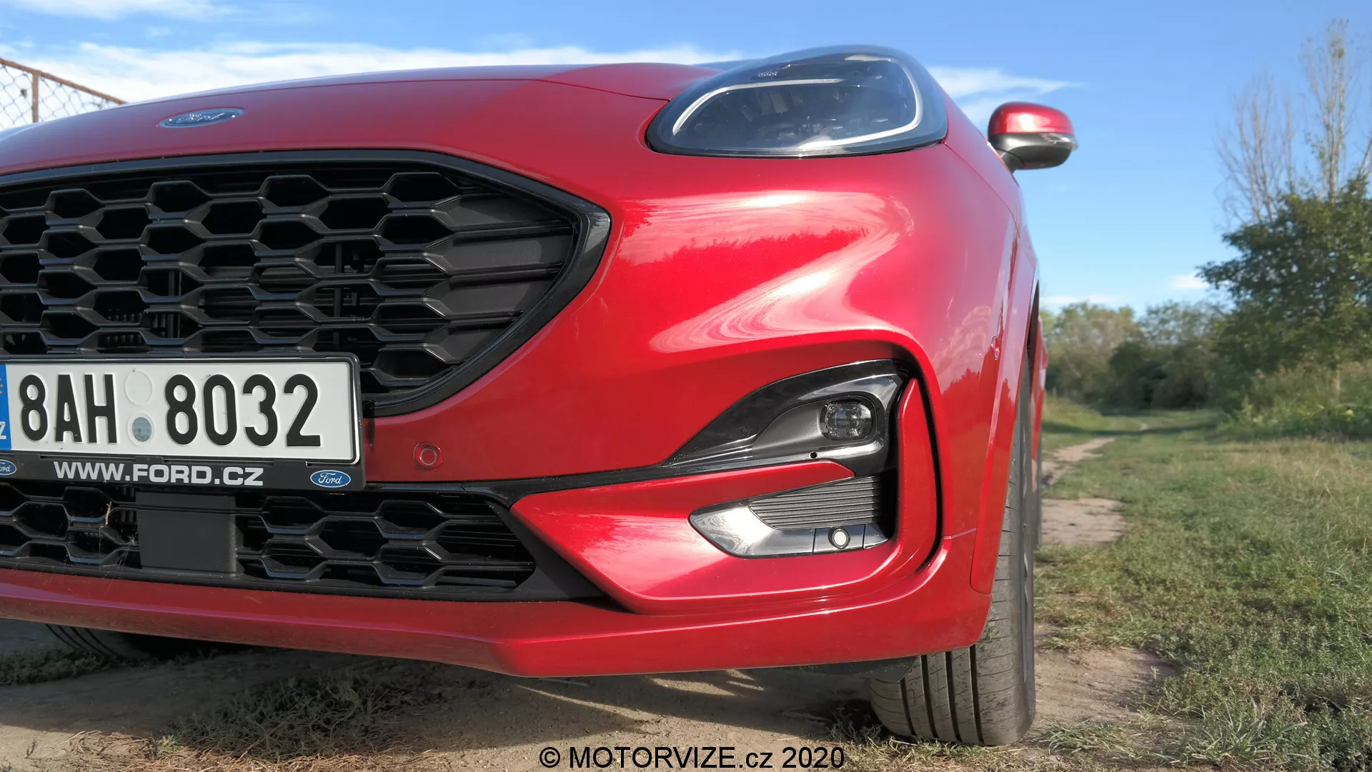 Primo piano angolato di una Ford Puma 2020 rossa con particolare attenzione al quarto anteriore sinistro. Caratteristiche includono griglia a nido d'ape, faro LED/HID elegante, paraurti anteriore e cofano aerodinamico scolpito. È visibile una targa europea con un sito web ceco. Elementi moderni come le frecce integrate nello specchietto laterale sono parzialmente visibili. L'immagine, ripresa da una bassa angolazione, utilizza luce naturale e bassa profondità di campo per mettere in risalto il design dell'auto in un ambiente esterno.