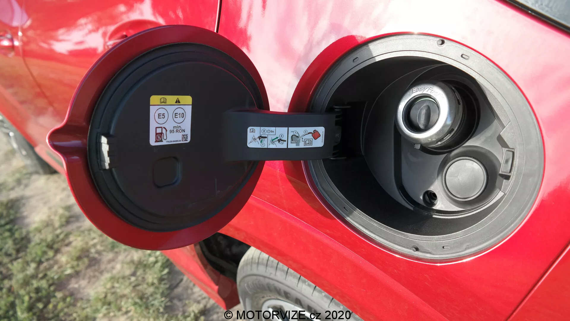 Vista ravvicinata di una presa di ricarica di Tipo 2 aperta su un veicolo elettrico o ibrido plug-in di colore rosso vibrante, con un focus sull'area della presa lucida, lo sportello incernierato e un'etichetta informativa visibile a sinistra all'interno della porta della presa, che suggerisce un'auto ben mantenuta con tecnologia avanzata per veicoli elettrici all'esterno.