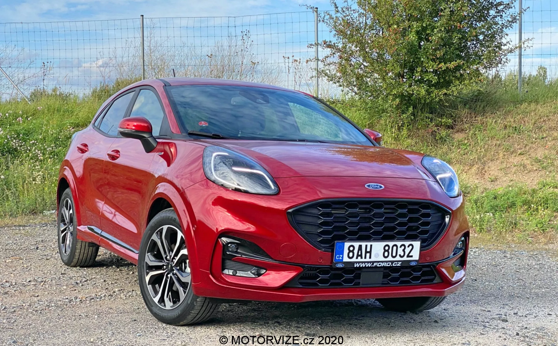L'immagine mostra una Ford Puma 2020 di colore rosso vivace, un veicolo crossover catturato da una vista frontale a tre quarti, che mette in evidenza la sua elegante griglia frontale a nido d'ape, fari angolari, cerchi in lega multispicchio, paraurti anteriore con fendinebbia integrati e linea delle spalle pronunciata. È visibile il cofano dinamico con pieghe, specchietti laterali con indicatori di direzione e parte delle porte anteriori e posteriori, indicando una configurazione a quattro o cinque porte, con uno sfondo di ghiaia e vegetazione.
