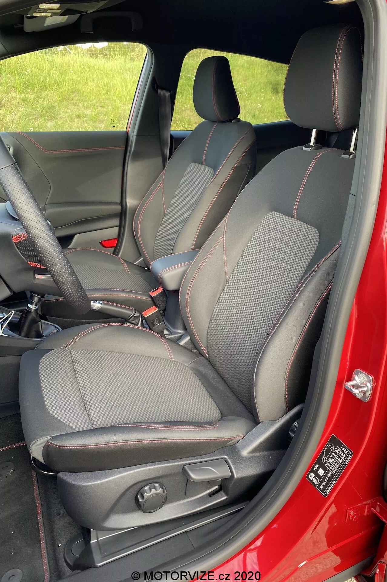 Immagine verticale dell'interno della Ford Puma 2020, focalizzata sul lato passeggero anteriore. Scattata dall'esterno con la porta aperta, mostra completamente il sedile del passeggero anteriore, parti del sedile del conducente, la console centrale e l'interno della porta. Il sedile ha un design sportivo con una combinazione di pelle e tessuto, cuciture a contrasto rosse, poggiatesta integrato e supporti laterali. I comandi manuali del sedile sono visibili. Il colore rosso dell'esterno dell'auto si riflette sul bordo della porta e il pannello interno della porta è nero. Sul telaio della porta c'è un'etichetta di sicurezza e informazioni.