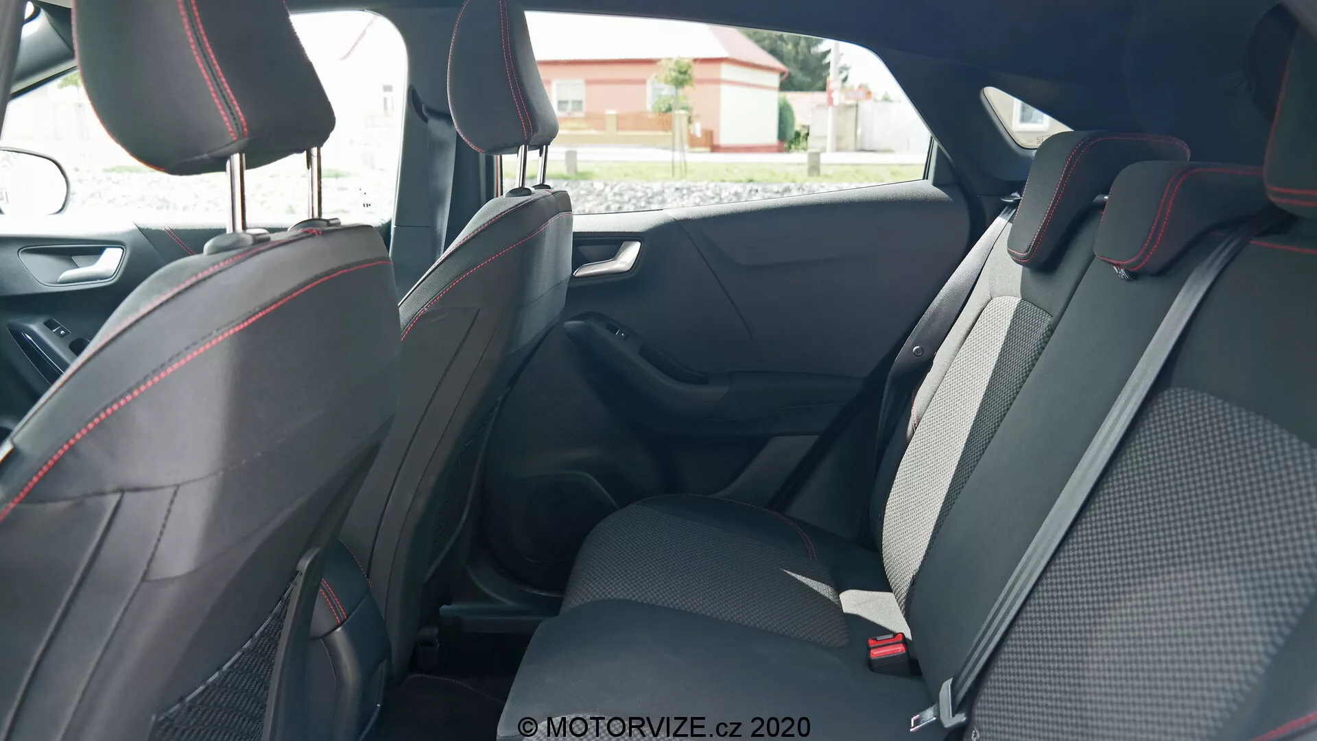 Scatto dell'interno della Ford Puma 2020 incentrato sui sedili posteriori, preso dalla porta posteriore del passeggero esterna, che mostra un design spazioso e sportivo con materiali in tessuto e pelle, accentuato da cuciture rosse. Le caratteristiche visibili includono poggiatesta regolabili, cinture di sicurezza a tre punti e sedili abbattibili per una maggiore versatilità. I pannelli delle porte con maniglie, comandi dei finestrini elettrici e spazi di stivaggio sono anche visibili, evidenziando aspetti ergonomici e di convenienza.