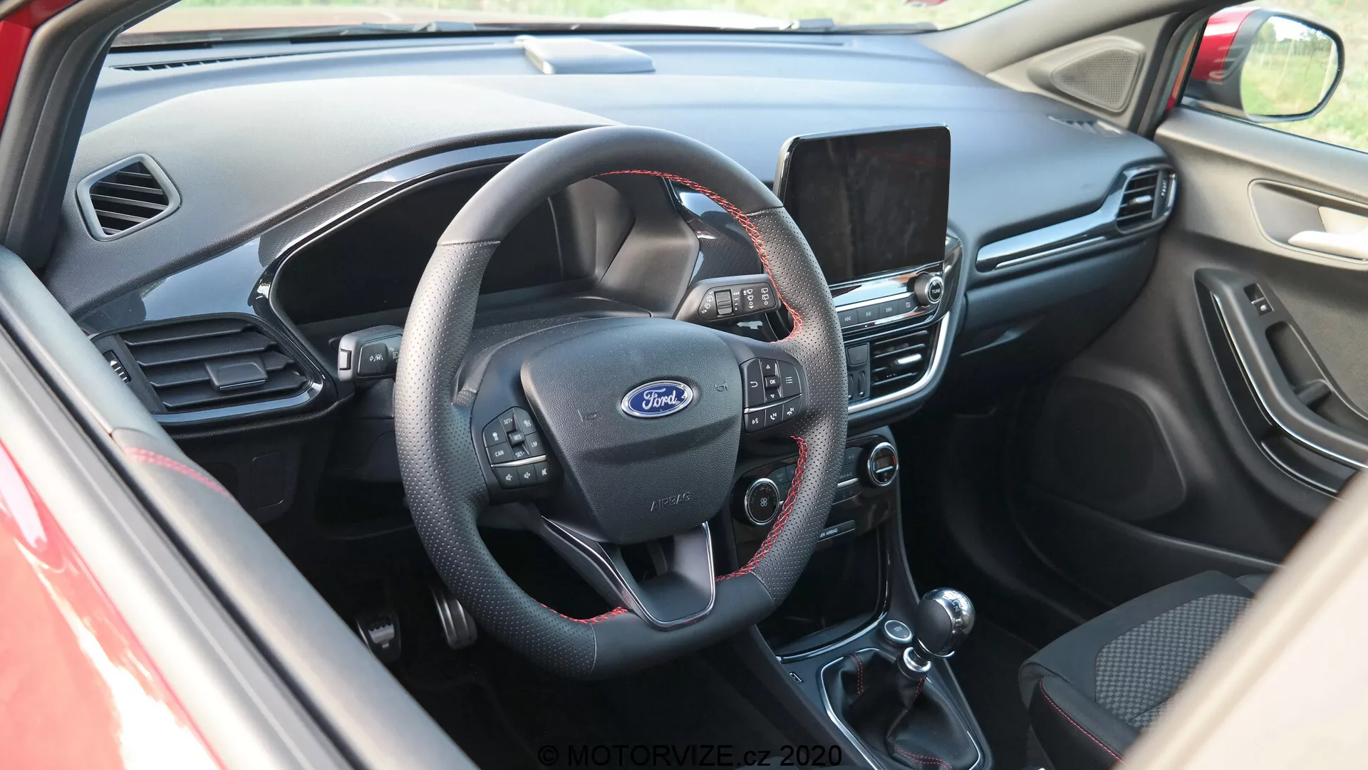 Vista interna del cockpit della Ford Puma 2020 da un'angolazione leggermente elevata, con un focus sul centro del cruscotto. Vista dalla porta del conducente, l'immagine mostra un volante a tre razze con pulsanti multifunzione per audio, comandi vocali e cruise control. L'emblema Ford è visibile al centro del volante. Dietro il volante è visibile una parte del quadro strumenti. Il cruscotto centrale presenta uno schermo di infotainment che probabilmente offre navigazione, riproduzione multimediale e impostazioni. Sotto di esso, si trova il sistema di controllo climatico manuale con quadranti rotativi e pulsanti per regolazione della temperatura, aria condizionata e sbrinamento. L'immagine rivela anche la leva del cambio automatico e la leva del freno a mano. L'interno combina plastica scura nella parte superiore del cruscotto con tessuto o pelle nera nelle parti inferiori, presentando un design funzionale focalizzato sulla praticità.