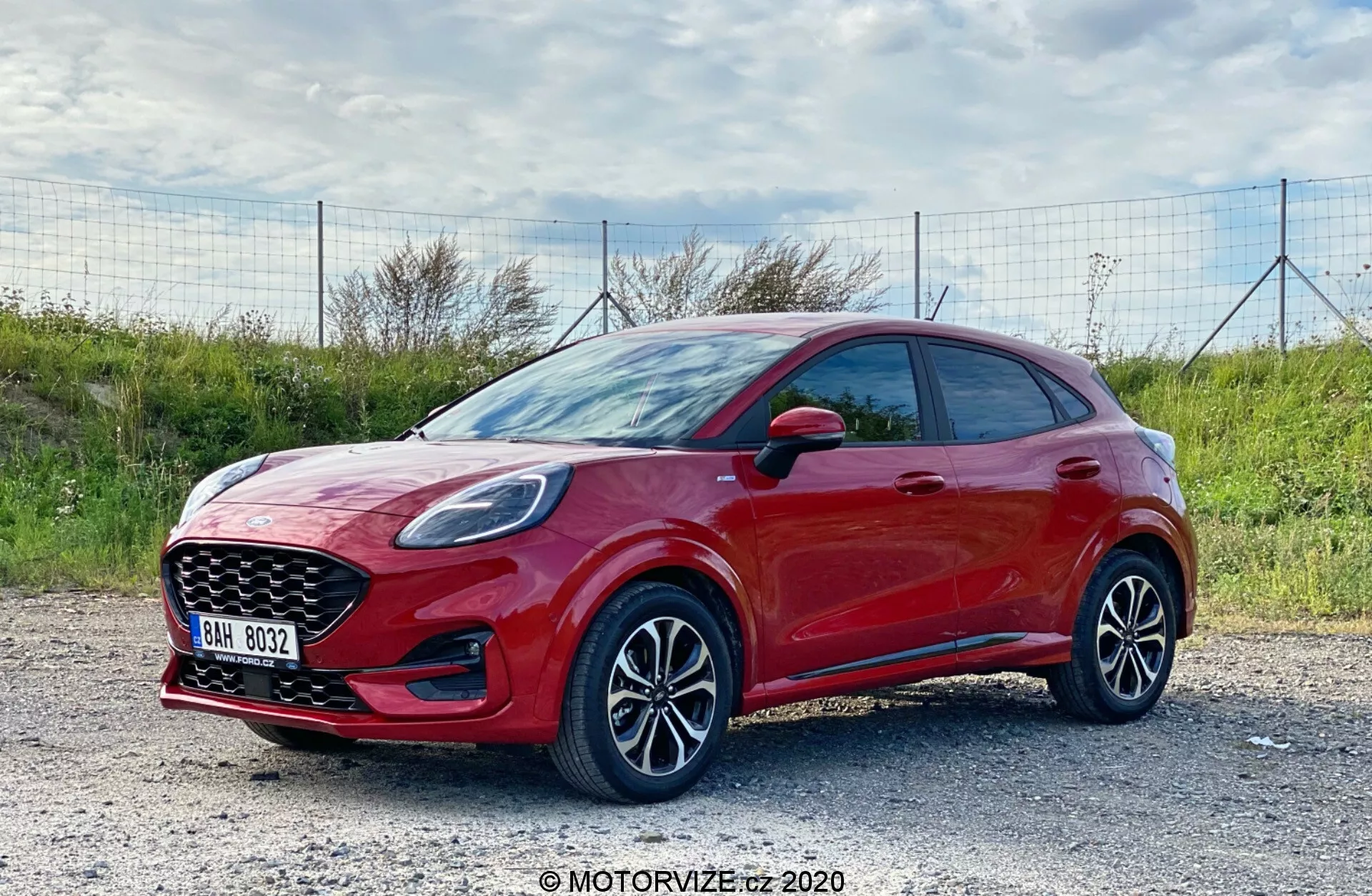 Immagine di profilo laterale di una moderna hatchback Ford Puma 2020, catturata alla luce del giorno con uno sfondo esterno naturale. Il veicolo di colore rosso vibrante con una finitura lucida è visto da un'angolazione di tre quarti anteriore, mostrando chiaramente il profilo laterale e la fascia frontale. L'auto è parcheggiata su una superficie ghiaiosa. La ripresa, effettuata da leggermente sopra il livello della ruota, mette in risalto i cerchi in lega a più razze e la parte inferiore del veicolo. Sono visibili i fari eleganti con probabile tecnologia LED, una griglia anteriore pronunciata e specchietti laterali con indicatori integrati. L'auto presenta maniglie delle porte del colore della carrozzeria, senza modanature protettive laterali visibili e uno stile sportivo hatchback con una linea del tetto discendente. Le caratteristiche tecnologiche potrebbero includere sistemi avanzati di assistenza alla guida e infotainment con opzioni di connettività.
