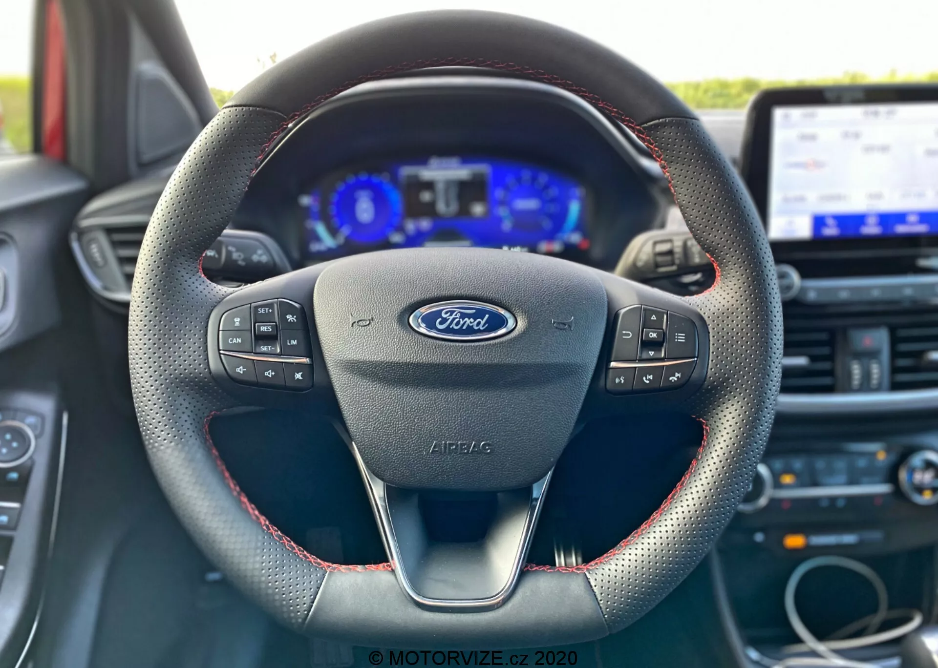 Scatto degli interni di una Ford Puma 2020 dalla prospettiva del sedile del conducente, focalizzato sul volante con logo Ford e comandi multifunzione, design moderno a tre razze con cuciture rosse che suggeriscono materiale in pelle, un quadro strumenti digitale affiancato da indicatori analogici e un sistema di infotainment touchscreen sullo sfondo.