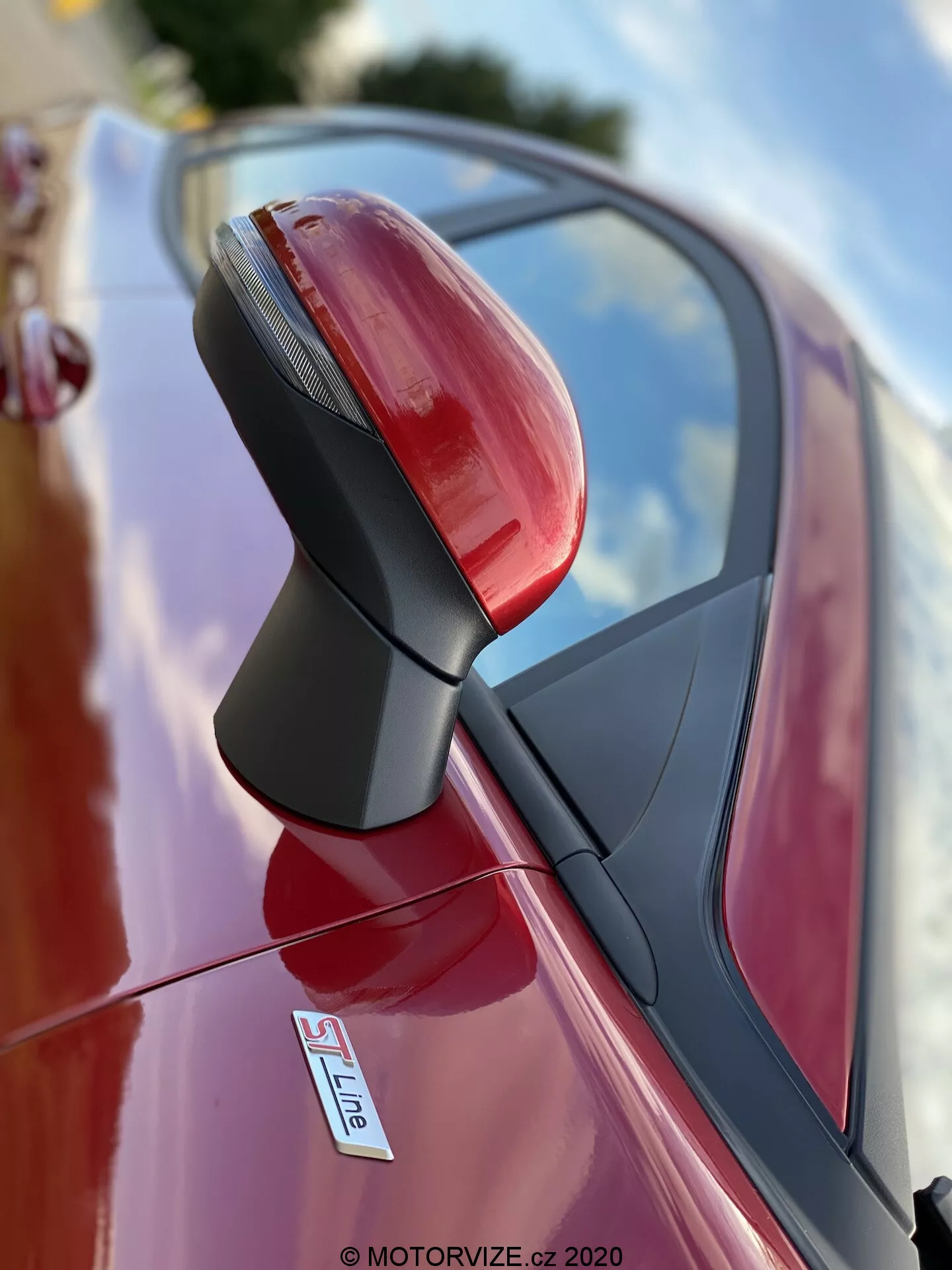 Primo piano dello specchietto retrovisore esterno lato guida di un'auto rossa, probabilmente un'Audi con distintivo S Line. La fotografia, scattata da un'angolazione bassa, si concentra sul gruppo specchio con una profondità di campo ridotta, sfocando lo sfondo. Lo specchio è nitido e dettagliato, mostrando indicatori di direzione integrati, un supporto nero strutturato e una finitura metallica lucida. I riflessi del cielo e dell'ambiente circostante indicano che l'auto è parcheggiata all'esterno.