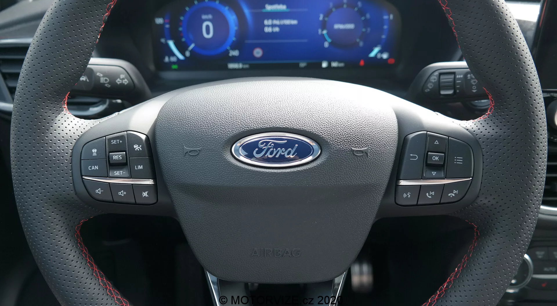 L'interno di una Ford Puma 2020 dalla prospettiva del conducente che mostra un volante sportivo in pelle con cuciture rosse, il logo centrale Ford, vari pulsanti di controllo, un cluster di strumenti digitale con velocimetro e tachimetro, e palette del cambio metalliche.