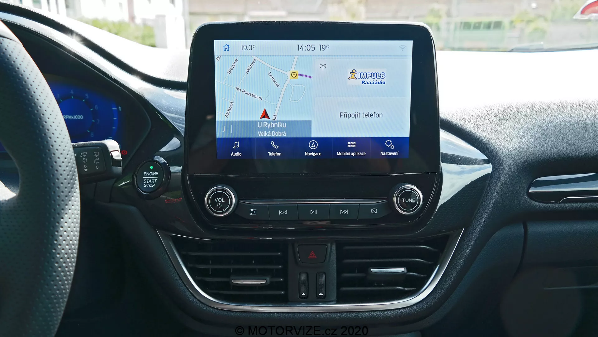 Consolle centrale dell'interno della Ford Puma 2020 dalla prospettiva del conducente o del passeggero anteriore, che mostra il sistema di infotainment con display touchscreen che mostra la mappa di navigazione, pulsanti fisici per luci di emergenza, regolazioni del controllo del clima, slot per CD, porta USB e presa di corrente da 12V. La consolle include anche un termometro digitale che segna 17°C e un orologio che mostra 14:05, il tutto circondato da elementi di design moderno con linee curve e prese d'aria del condizionatore.