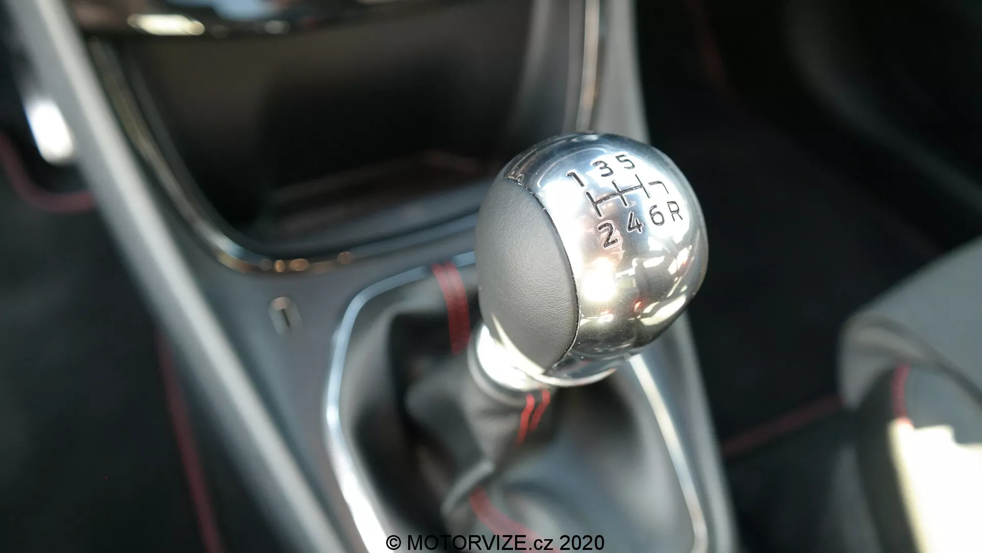 Fotografia ravvicinata della leva del cambio manuale della Ford Puma 2020, vista da un'angolazione leggermente elevata che mostra il profilo superiore e laterale. Il pomello del cambio metallico presenta una parte superiore nera con numeri bianchi incisi che indicano un modello di trasmissione a 6 marce con una 'R' per la retromarcia. La leva del cambio è montata su una base rifinita in cromo e coperta da un manicotto nero, concentrandosi nitidamente sul pomello del cambio mentre lo sfondo è sfocato, evidenziando l'interno del veicolo e la tecnologia della trasmissione.