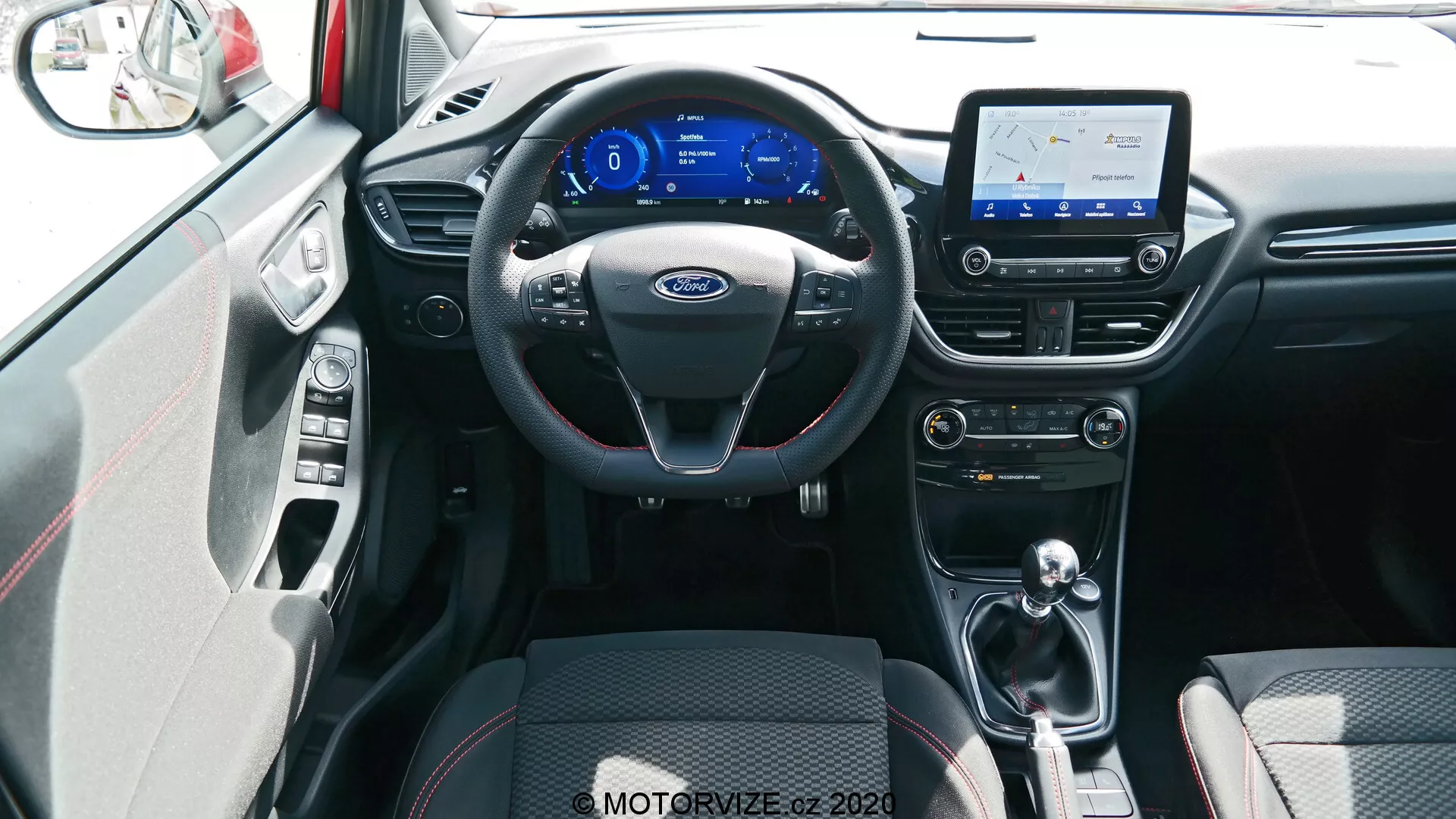 Vista interna della Ford Puma 2020, presa dalla posizione centrale dei sedili posteriori. L'immagine mostra un volante nero a tre razze con controlli multifunzione e accenti argentati, un cruscotto digitale con un display a tema blu, e uno schermo touch di infotainment che mostra una mappa, indicando capacità di navigazione. Sotto lo schermo ci sono i controlli per il condizionamento dell'aria e la ventilazione con manopole e pulsanti per regolare la temperatura e la velocità della ventola. Il tunnel centrale ospita una leva del cambio, suggerendo una trasmissione automatica o automatica manuale. I sedili anteriori presentano una tappezzeria in tessuto bicolore nero con motivi per presa e comfort, e una sezione del pannello interno della porta, inclusa una maniglia argentata della porta e accenti abbinati, è visibile. Il pavimento è coperto di tappeti che si fondono con lo schema dei colori interni e probabilmente c'è una buona illuminazione ambientale. La ripresa è fatta da un angolo leggermente elevato, mostrando in modo completo l'interno anteriore e le caratteristiche tecnologiche disponibili.