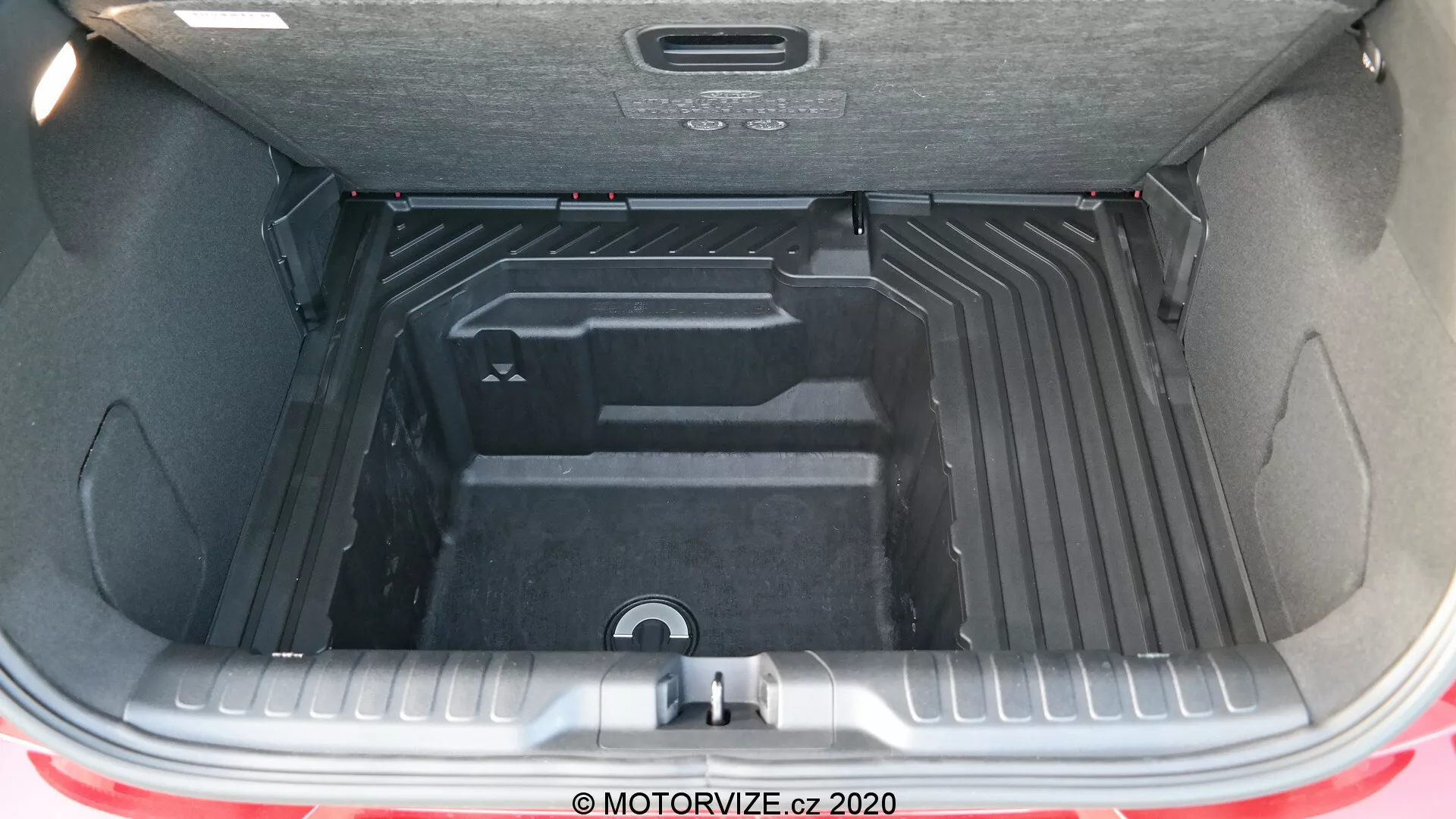 Una vista dettagliata del spazioso e versatile bagagliaio della Ford Puma 2020 con il portellone aperto, che mostra l'interno vuoto, pulito e ordinatamente sagomato progettato per l'organizzazione di bagagli e carico. L'interno del bagagliaio è realizzato con un materiale resistente e facile da pulire, con sezioni rialzate e incassate per soluzioni di stoccaggio ottimizzate, incluso una maniglia incassata centrale per l'accesso a un vano inferiore o al vano per la ruota di scorta. Sono parzialmente visibili il paraurti posteriore e una striscia del colore rosso della carrozzeria, insieme alla copertura isolante del portellone attrezzata con una maniglia per facilitare la chiusura.
