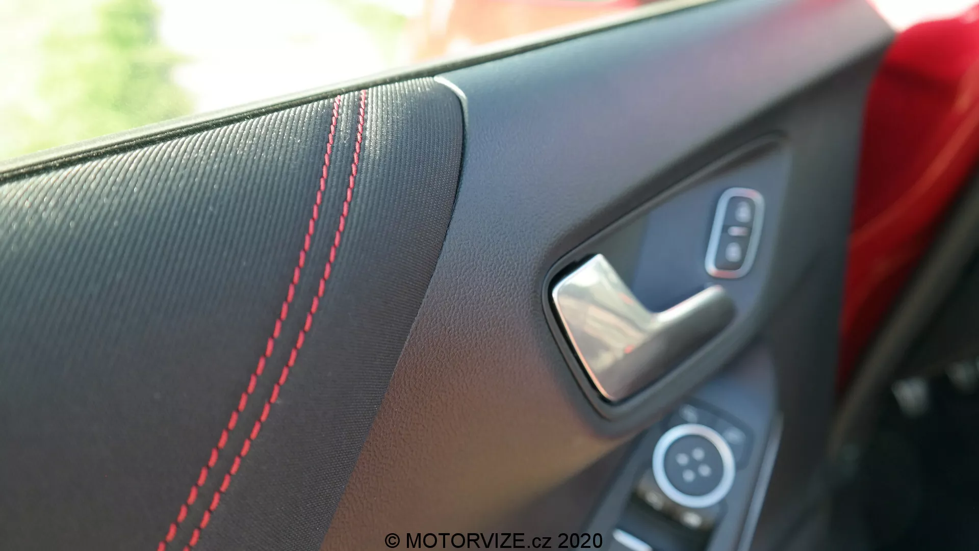 Vista ravvicinata del pannello porta interno della Ford Puma 2020 dalla prospettiva di un passeggero o del conducente all'interno del veicolo. L'immagine si concentra sulla zona della maniglia, mettendo in evidenza l'inserto testurizzato con cuciture a contrasto rosse, la maniglia della porta in metallo e una parte del pannello di controllo con l'interruttore del finestrino elettrico. La bassa profondità di campo sfoca lo sfondo, enfatizzando gli elementi di design sportivi e lussuosi in luce naturale diurna.