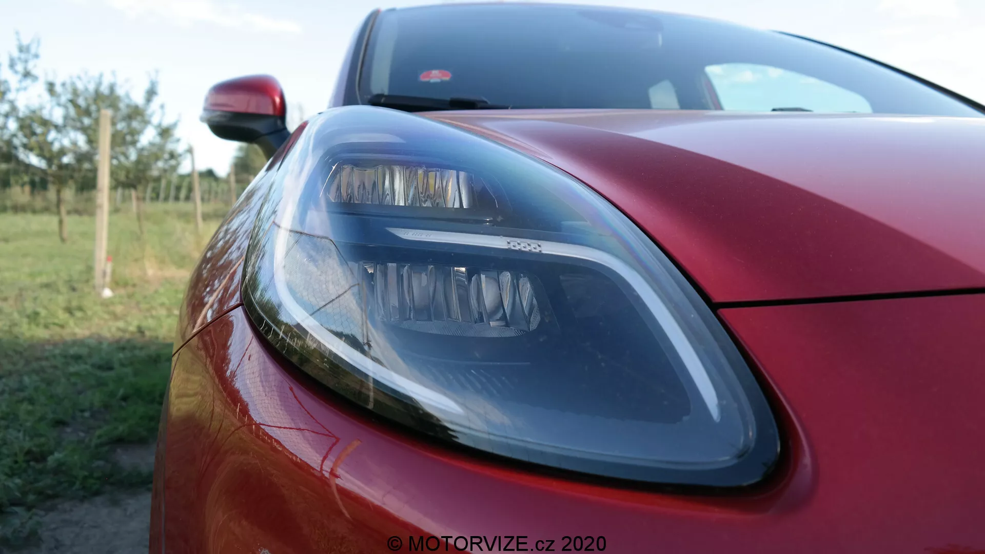 Primo piano della sezione anteriore sinistra di una Ford Puma 2020 rosso metallizzato, che mette in risalto un moderno faro con elementi di design eleganti, parte del cofano e uno scorcio della griglia anteriore, con una profondità di campo ridotta che sfoca lo sfondo, mettendo in evidenza la finitura esterna premium e la tecnologia di illuminazione.
